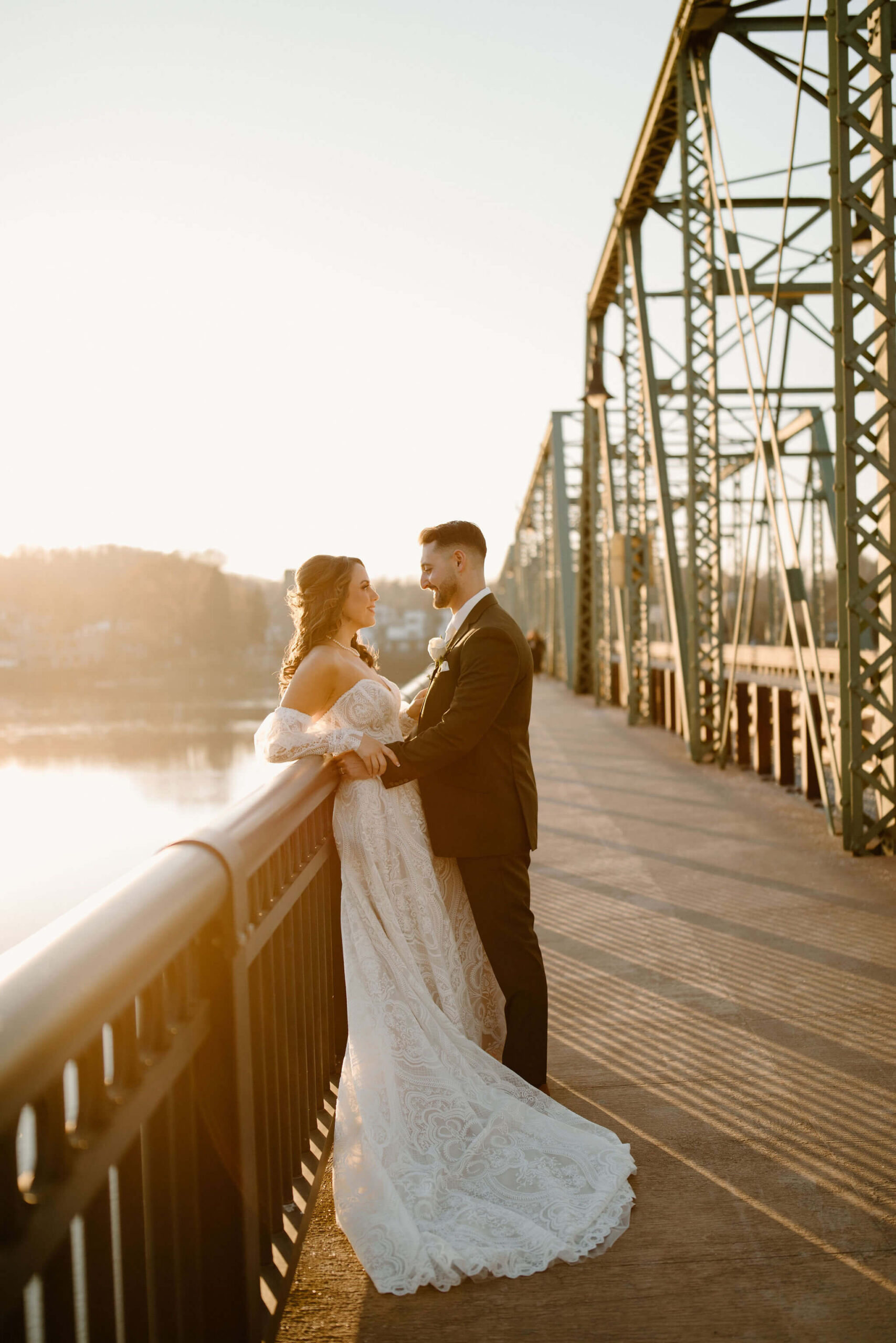 Bride (off-white boho dress) with her back against a railing of a bridge overlooking a river at sunset. The Groom has his arms around her waist and they are smiling at each other.