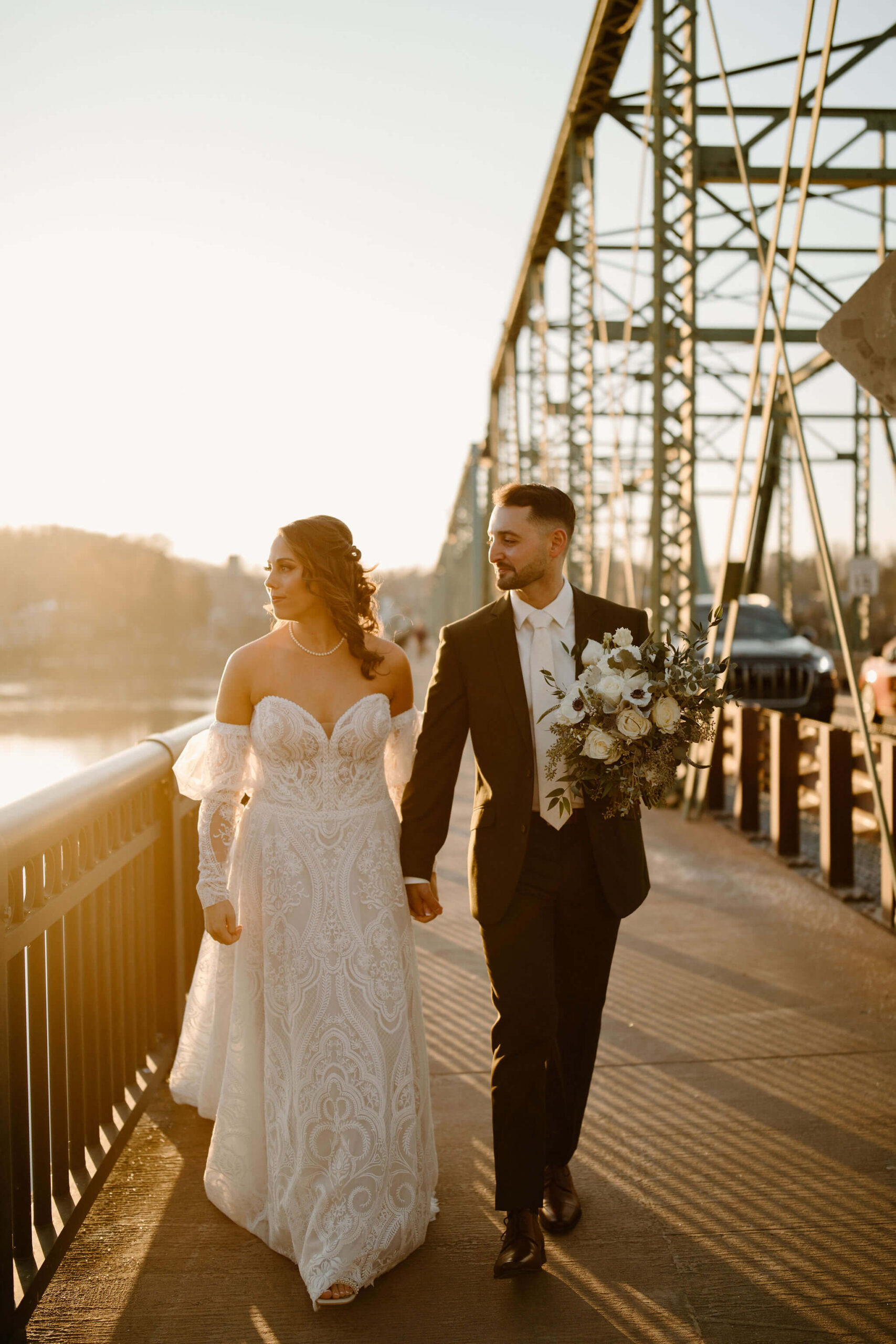 ride and groom walking on a bridge toward the camera. groom is holding the bridal flower bouquet. they are both looking to the left off-camera, toward a river.