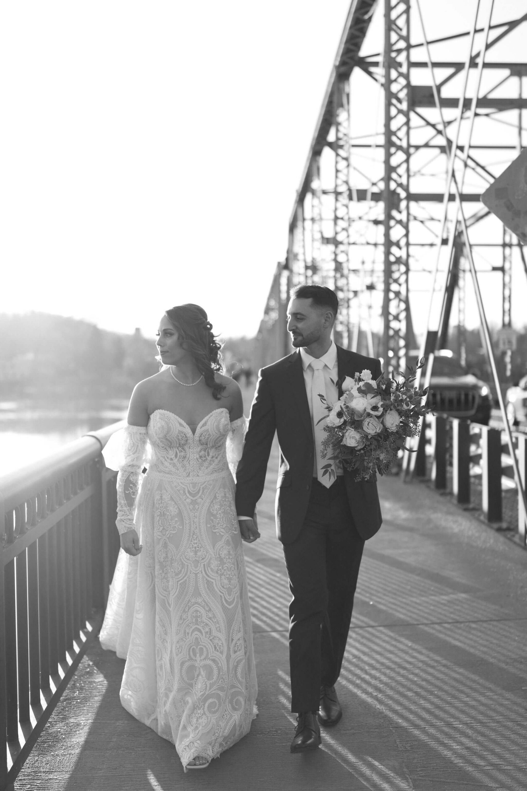 black and white image of bride and groom walking on a bridge toward the camera. groom is holding the bridal bouquet. they are both looking to the left off-camera, toward a river.