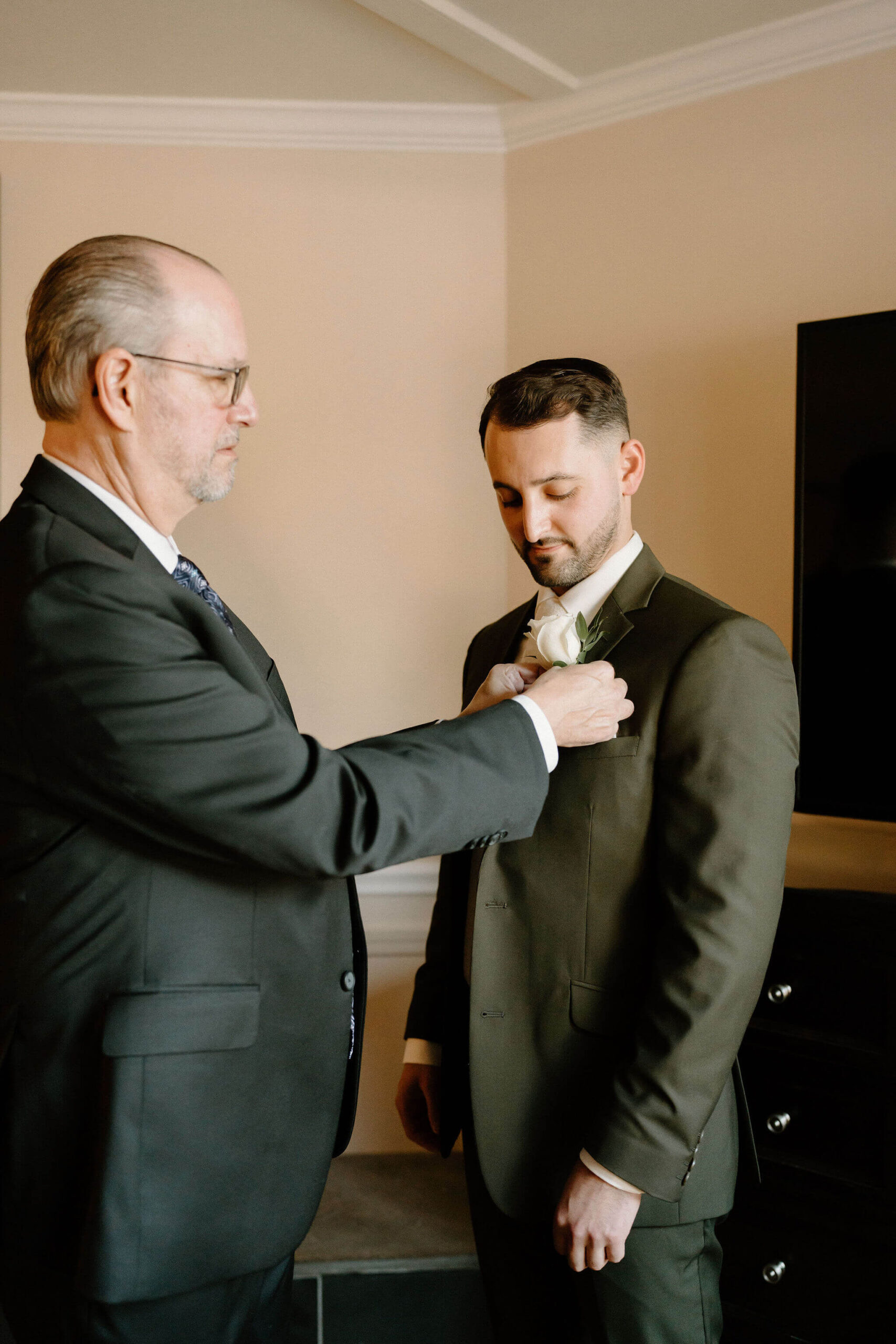 father of the groom (white slicked back hair and a black suit) helping the groom (brown hair and green suit) to pin his bouttoniere