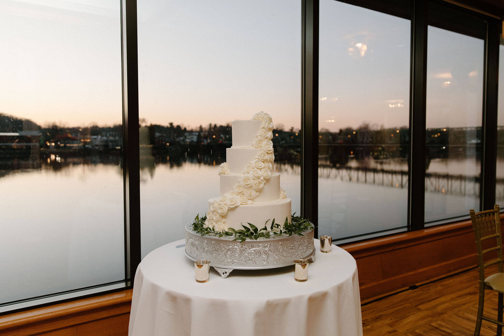 four-tiered white wedding cake, with a thick band of white roses wrapping around the cake from top-to-bottom, with the sun setting out the window behind it