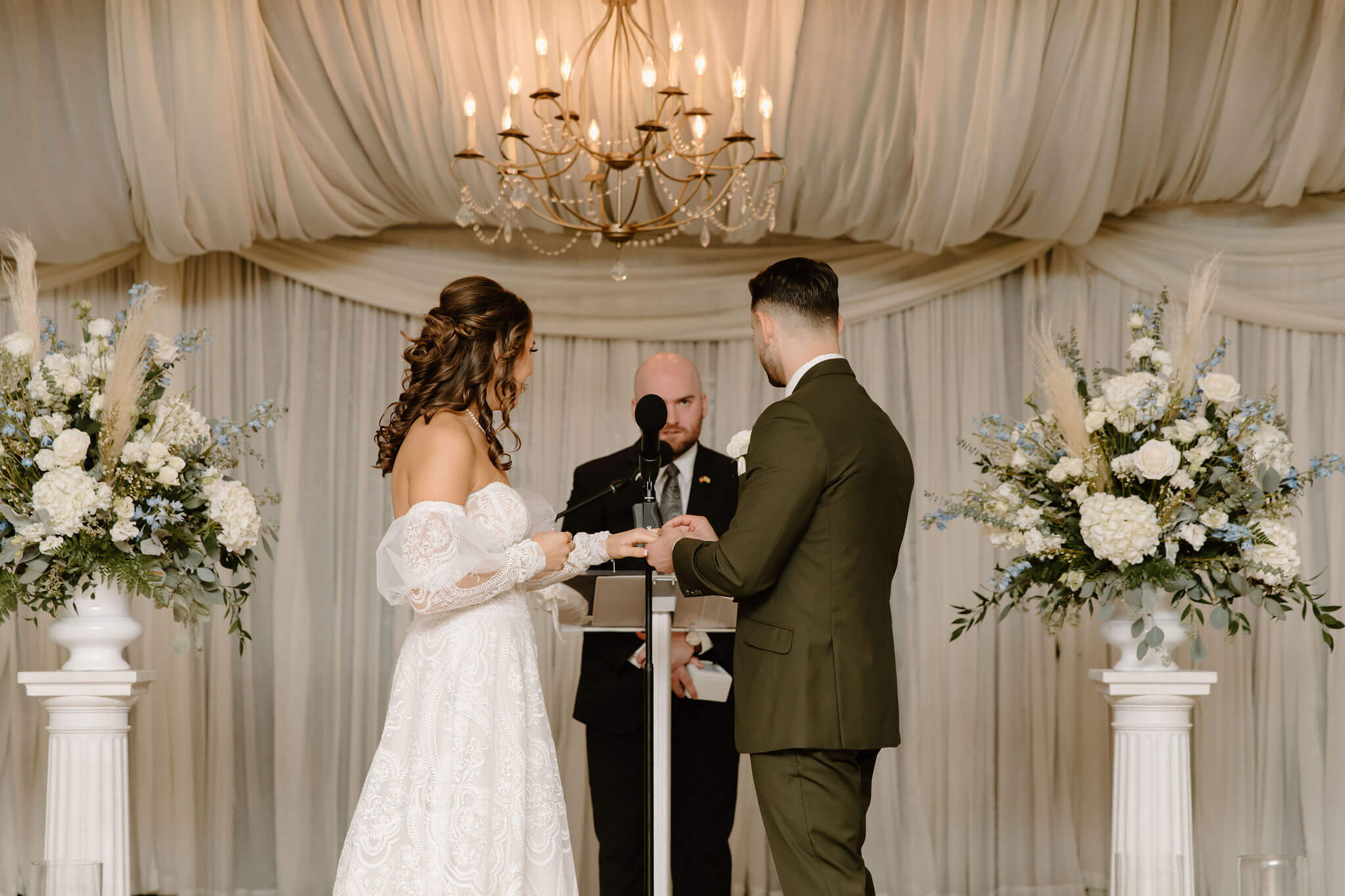 groom (green suit) putting a wedding band on his bride's (white dress) finger during their wedding ceremony