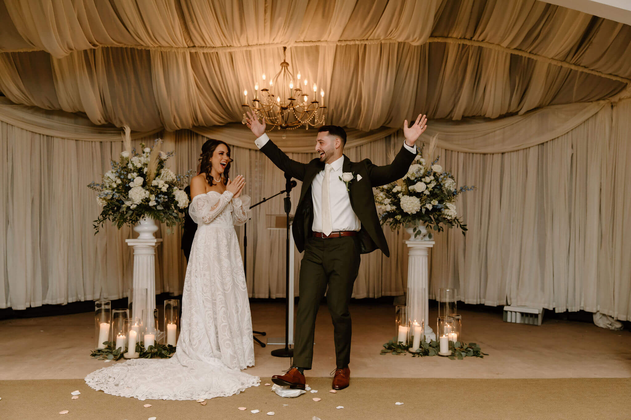 groom (green suit, hands lifted in celebration) and bride (white dress, clapping her hands and smiling) celebrating after breaking the glass in their Jewish wedding ceremony