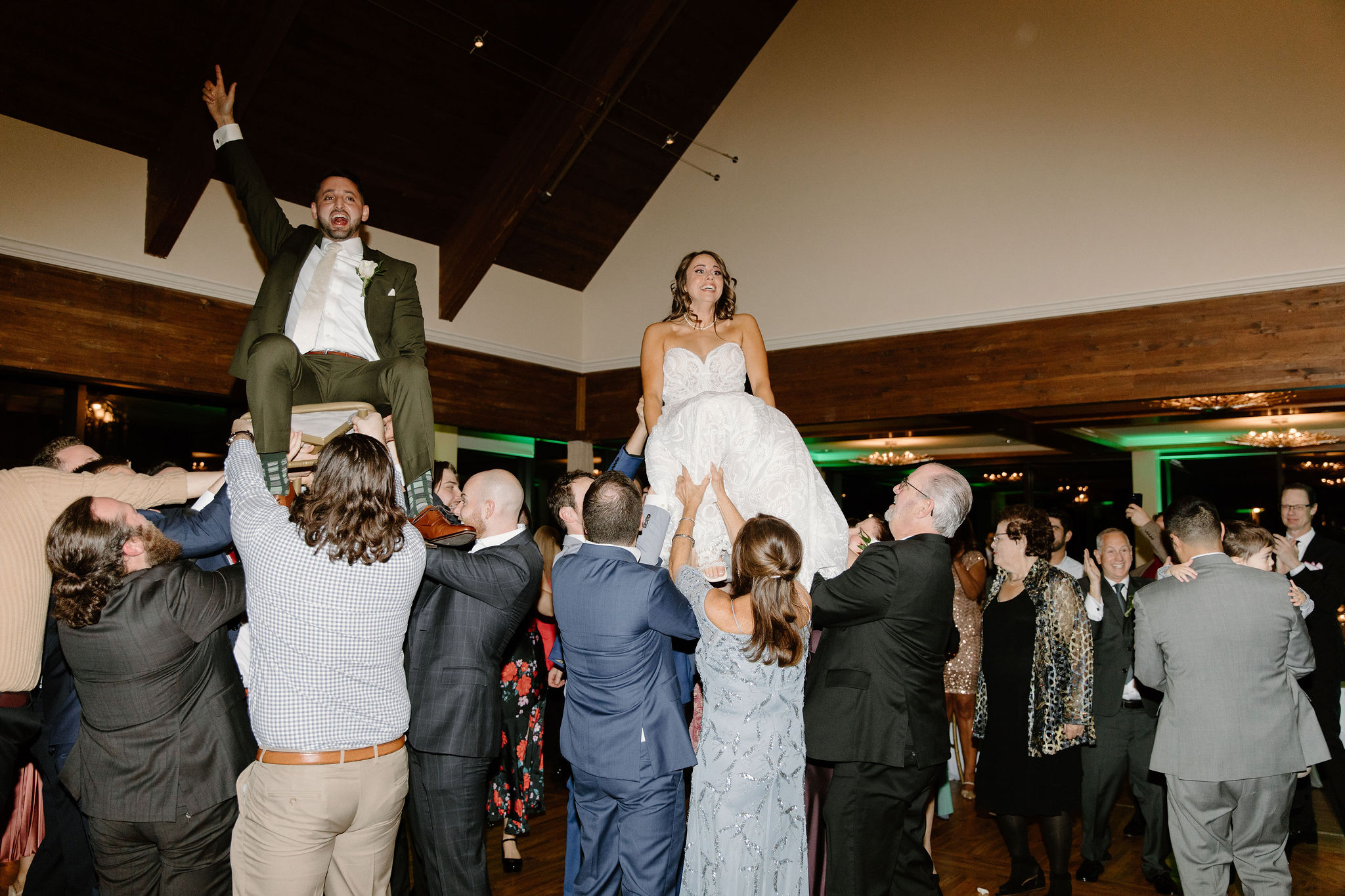 bride (white dress) and groom (green suit) being lifted in the air on chairs by their wedding guests in a traditional Jewish dance called the Hora