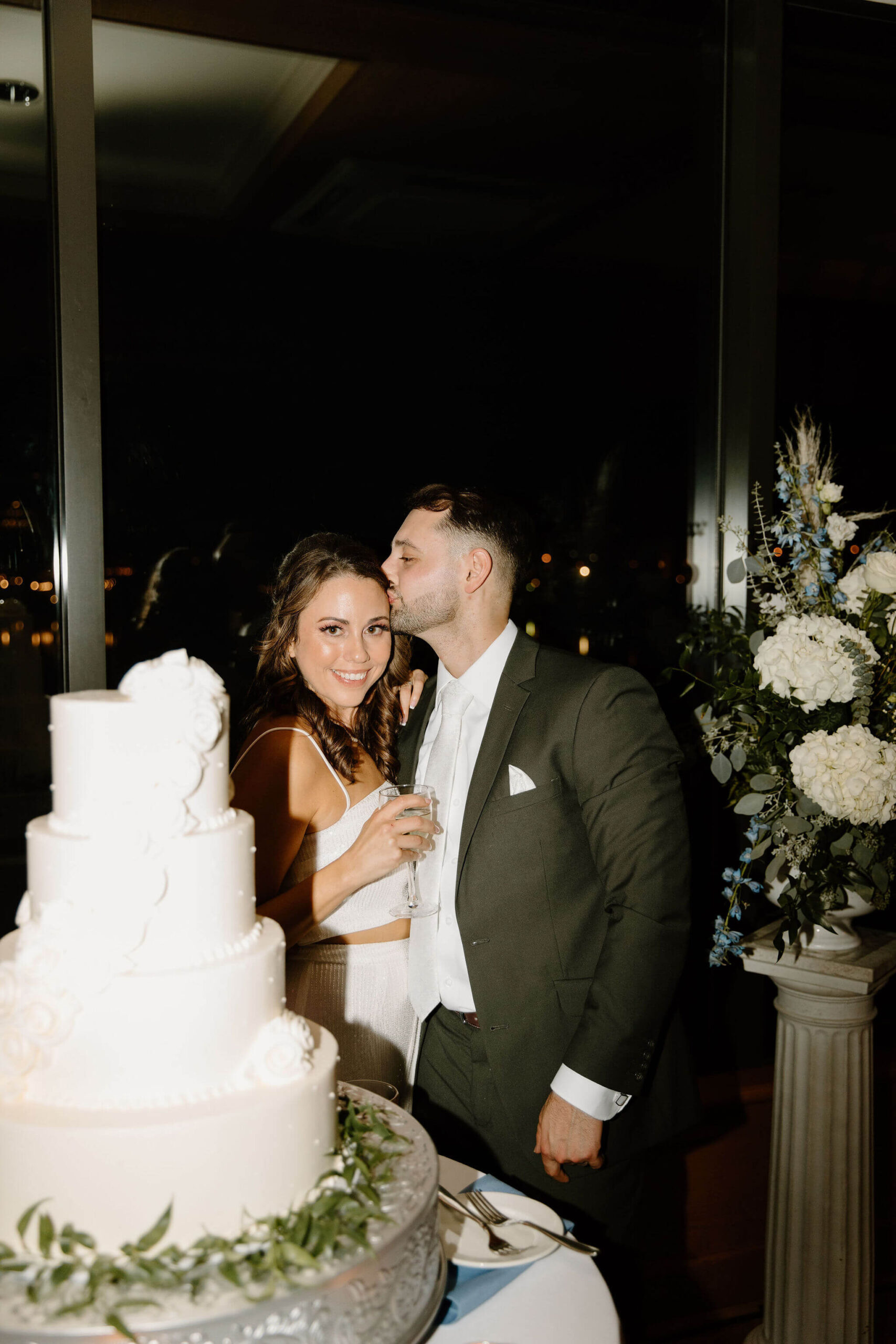 groom kissing bride on the cheek while bride smiles at the camera as they prepare to cut their wedding cake