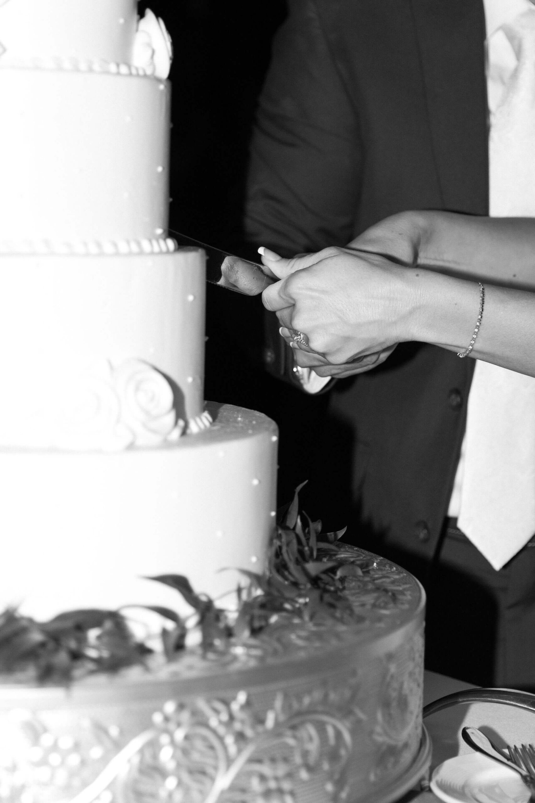 close up black and white image of bride and groom's hands cutting their four-tiered wedding cake