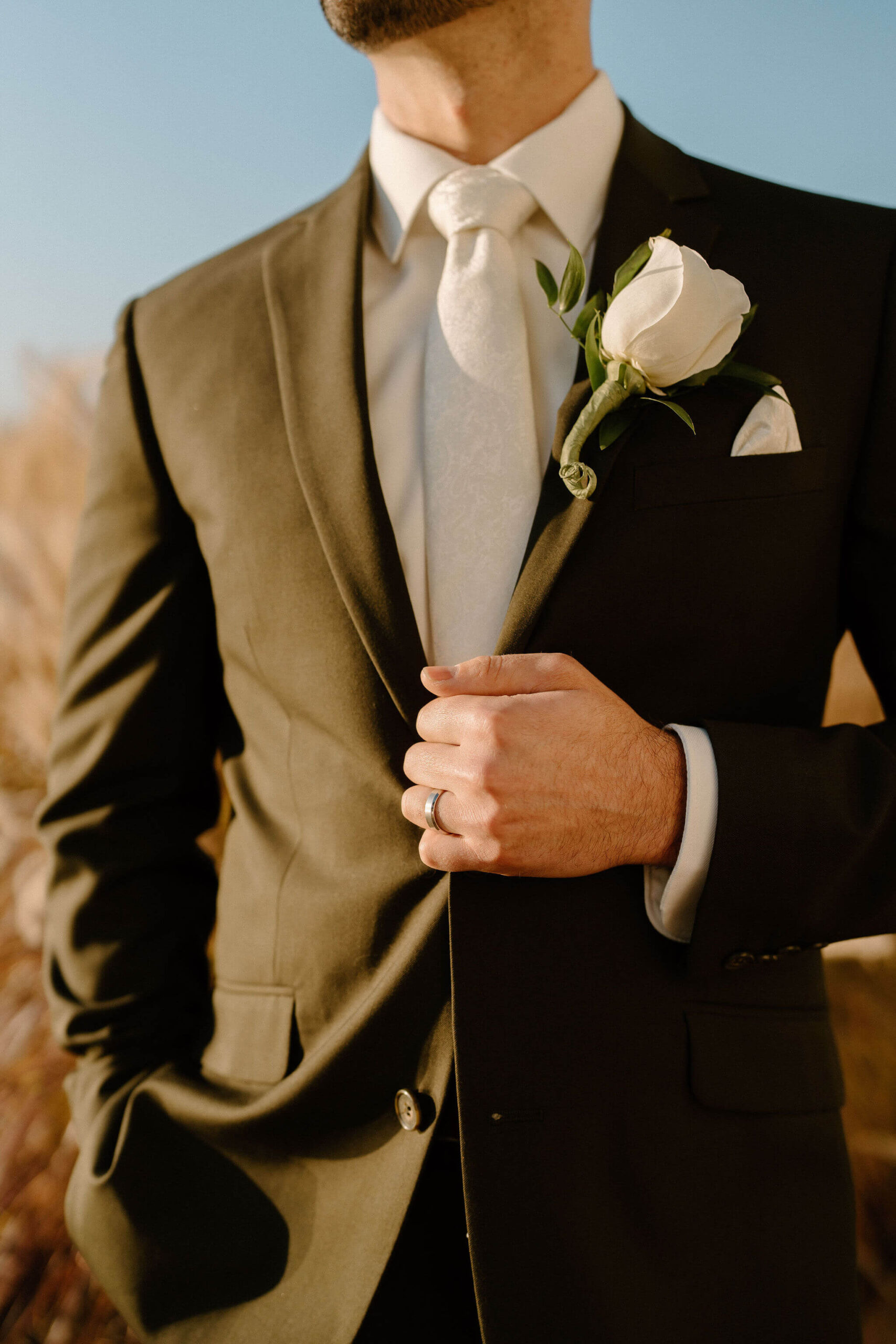 close up portrait of a groom (dark green suit, ivory tie, and white rose bouttonniere) holding his suit jacket with his left hand (and a silver wedding band)