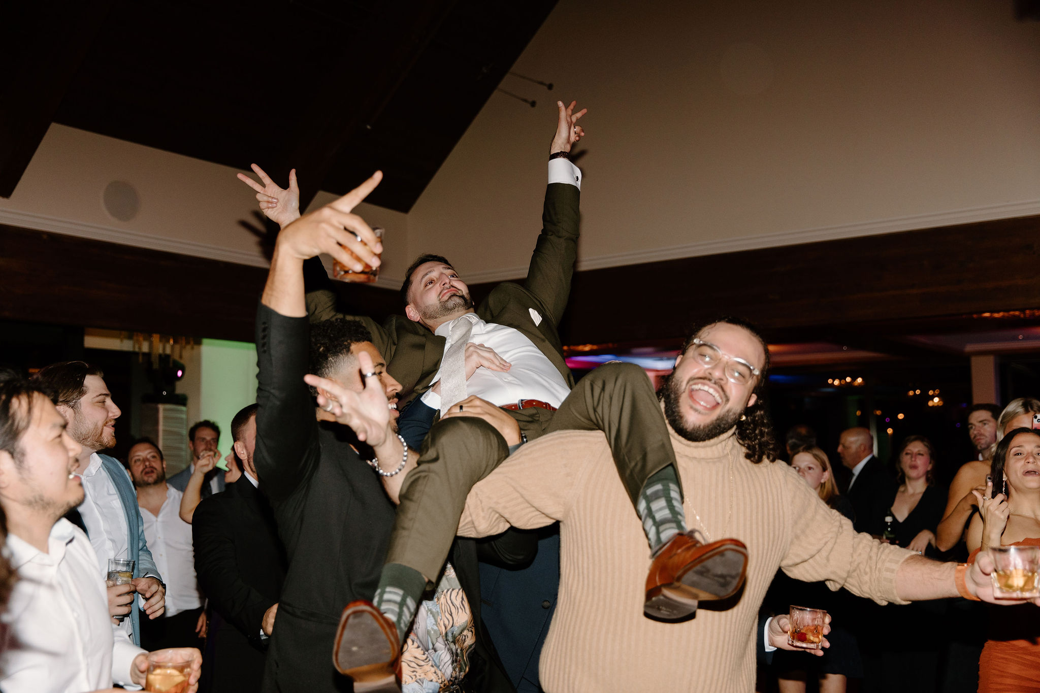 groom (green suit) waving his hands in the air as his friends lift him on their shoulders, all smiling and cheering