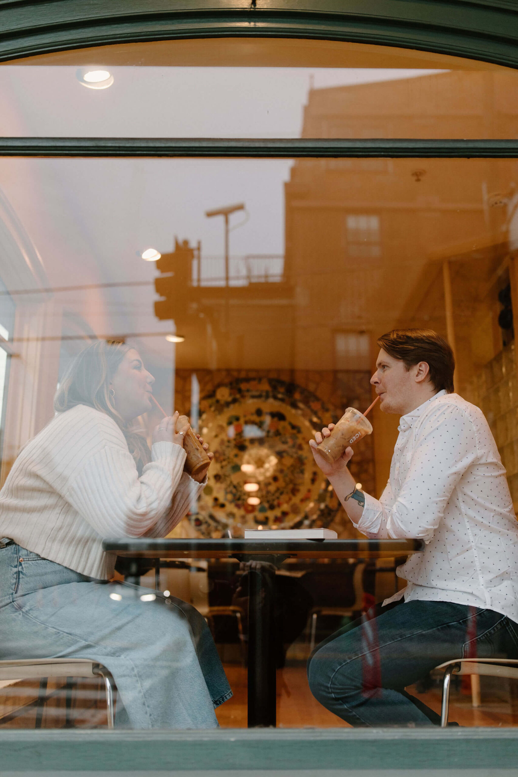 a man and a woman sitting across from each other at a table sipping iced coffees, photographed through a window from the street