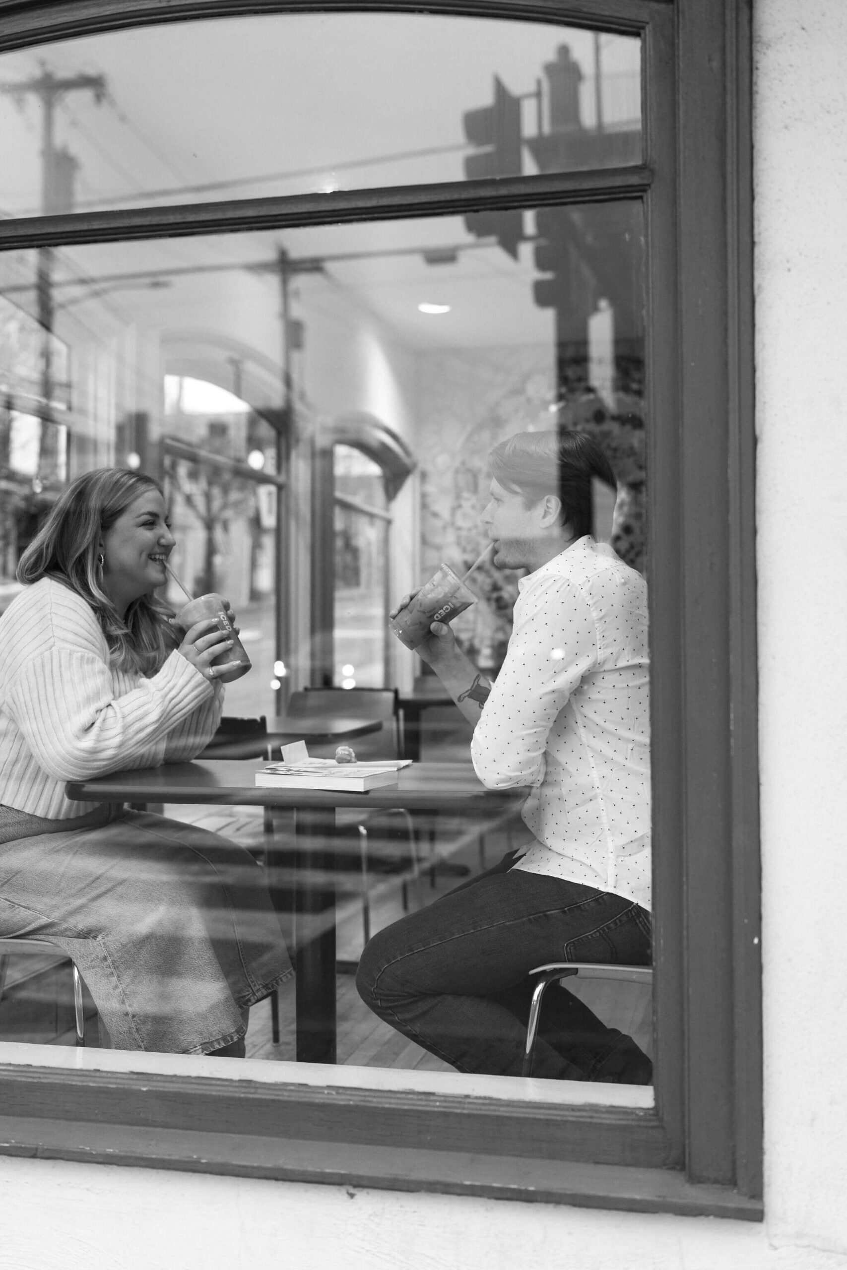 black and white image a man and a woman sitting across from each other at a table sipping iced coffees and smiling, photographed through a window from the street