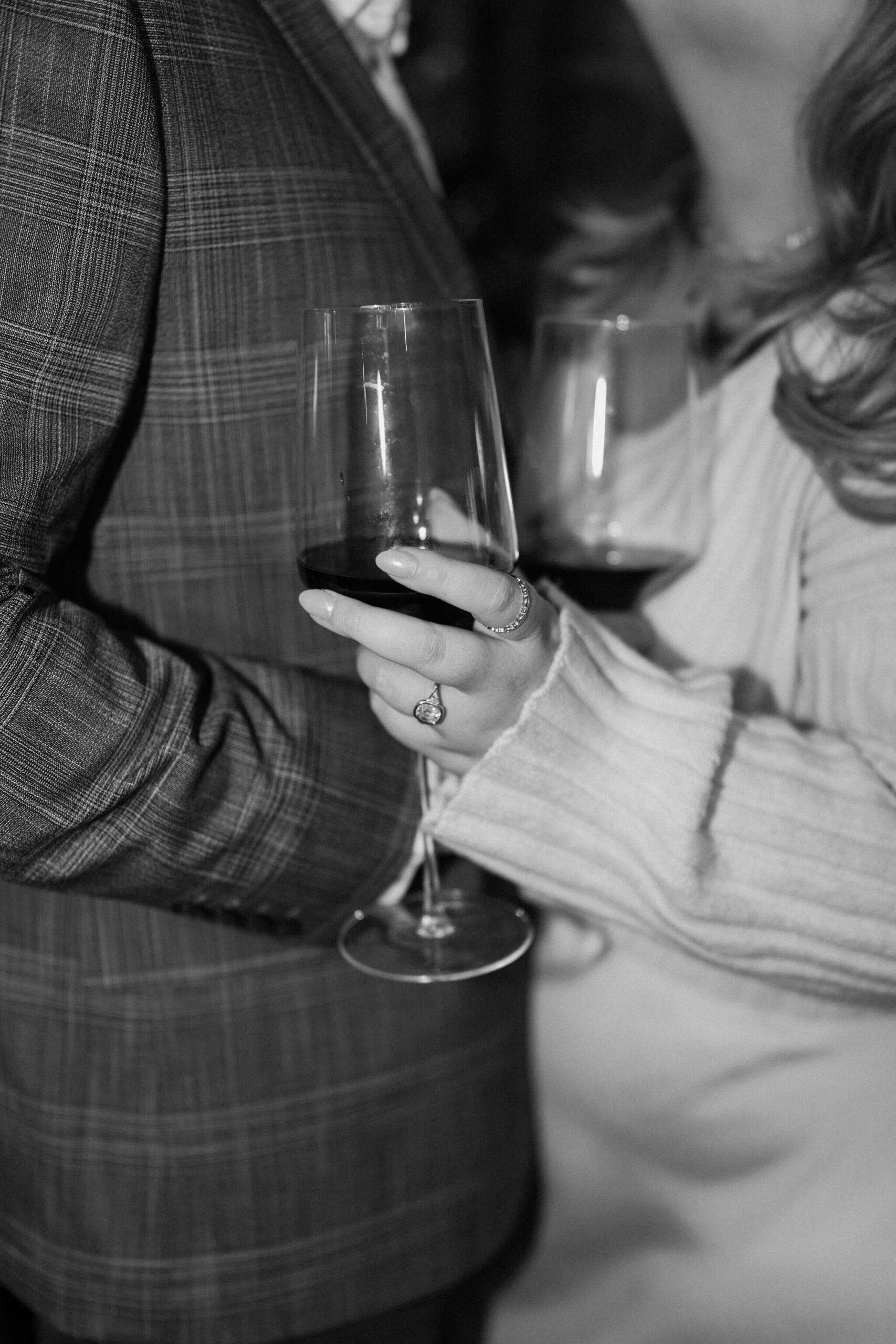 black and white close up image of a couple standing close together, both holding wine glasses, with an engagement ring on her left hand holding the glass