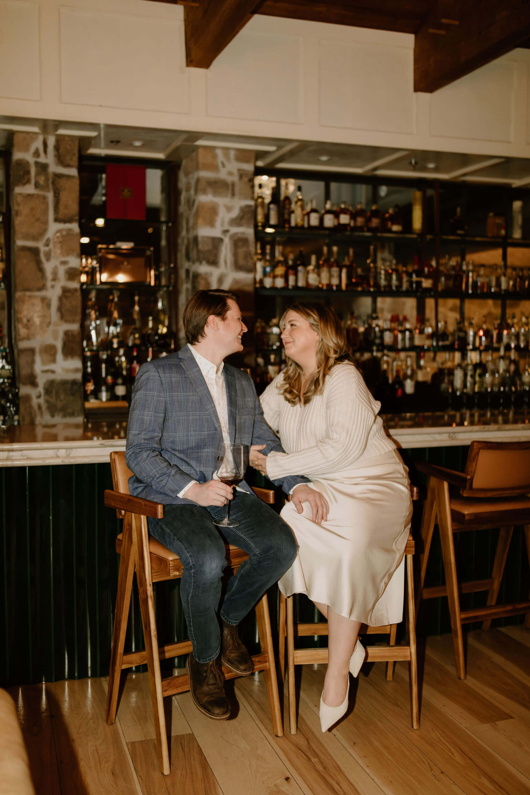 a couple seated at a bar (he is in a blue suit jacket, she is in a white sweater and skirt), leaning toward each other with his hand on her lef