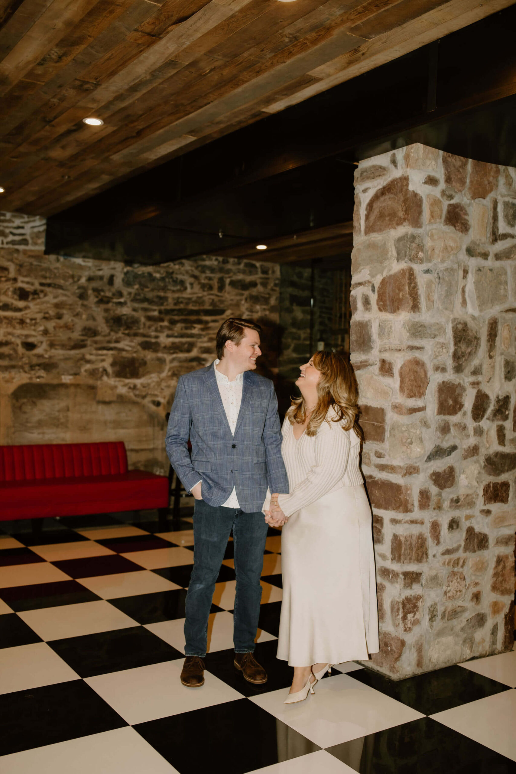 man (light brown hair, blue plaid suit jacket, jeans, and boots) holding hands with a woman (long blonde hair, white sweater, white skirt), looking at each other and smilng against a stone wall and black and white checkered floor