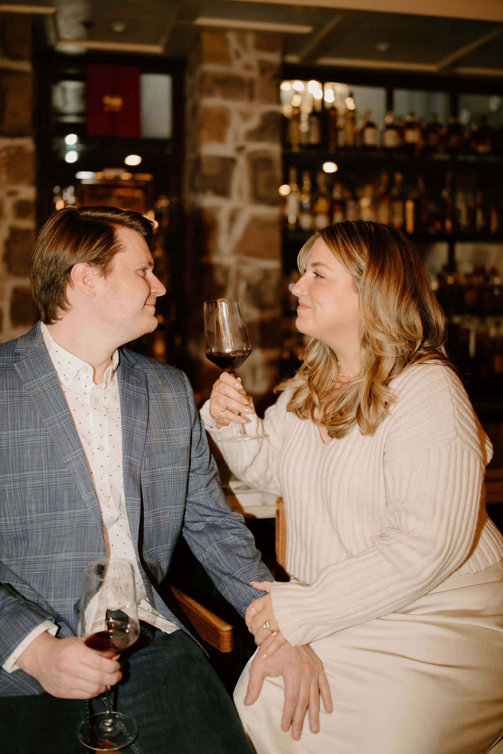 man and a woman seated at a bar, both holding wine glasses, with his hand on her knee, smiling softly at each other