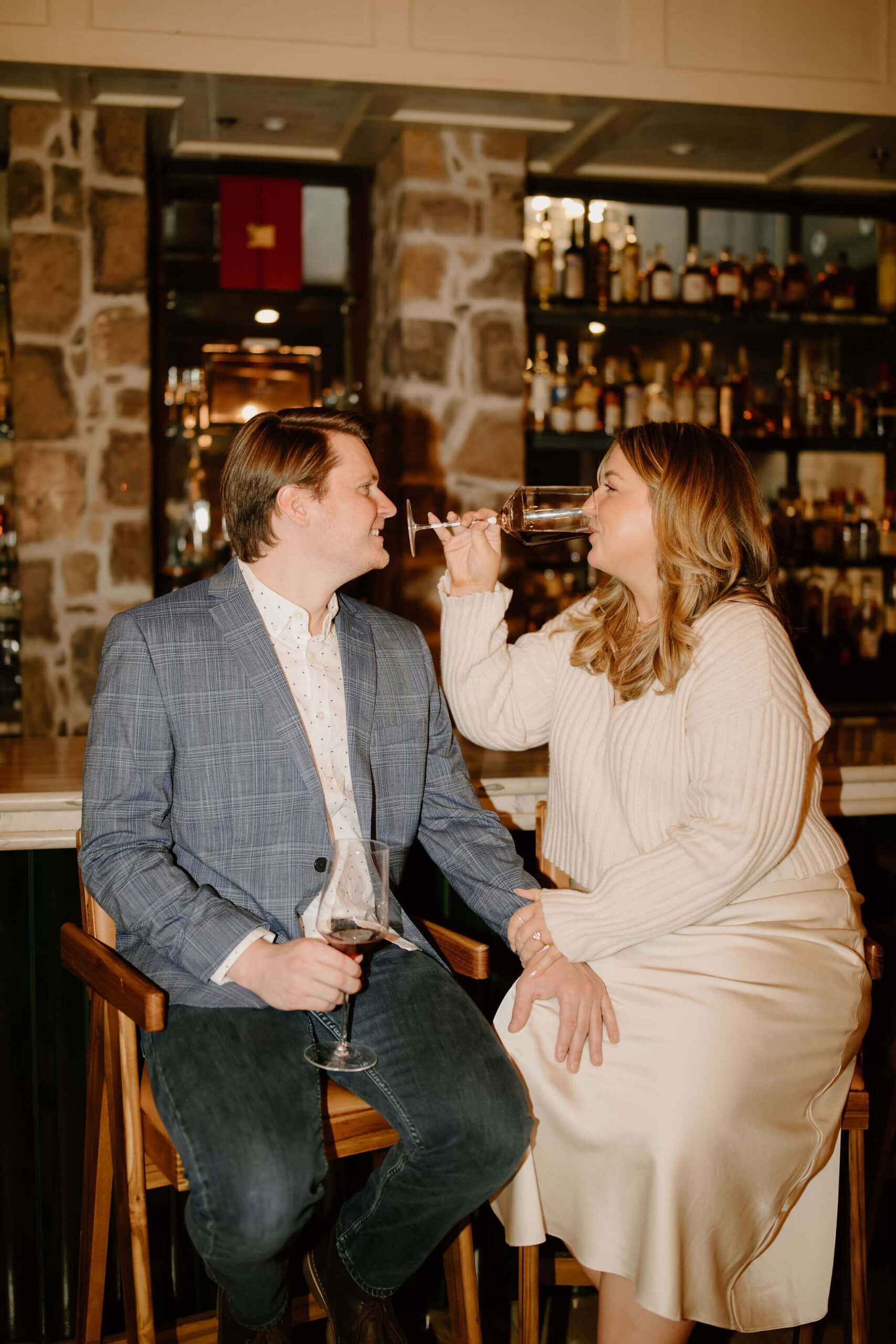 man and a woman seated at a bar, both holding wine glasses, with his hand on her knee, smiling softly at each other while she takes a sip of wine