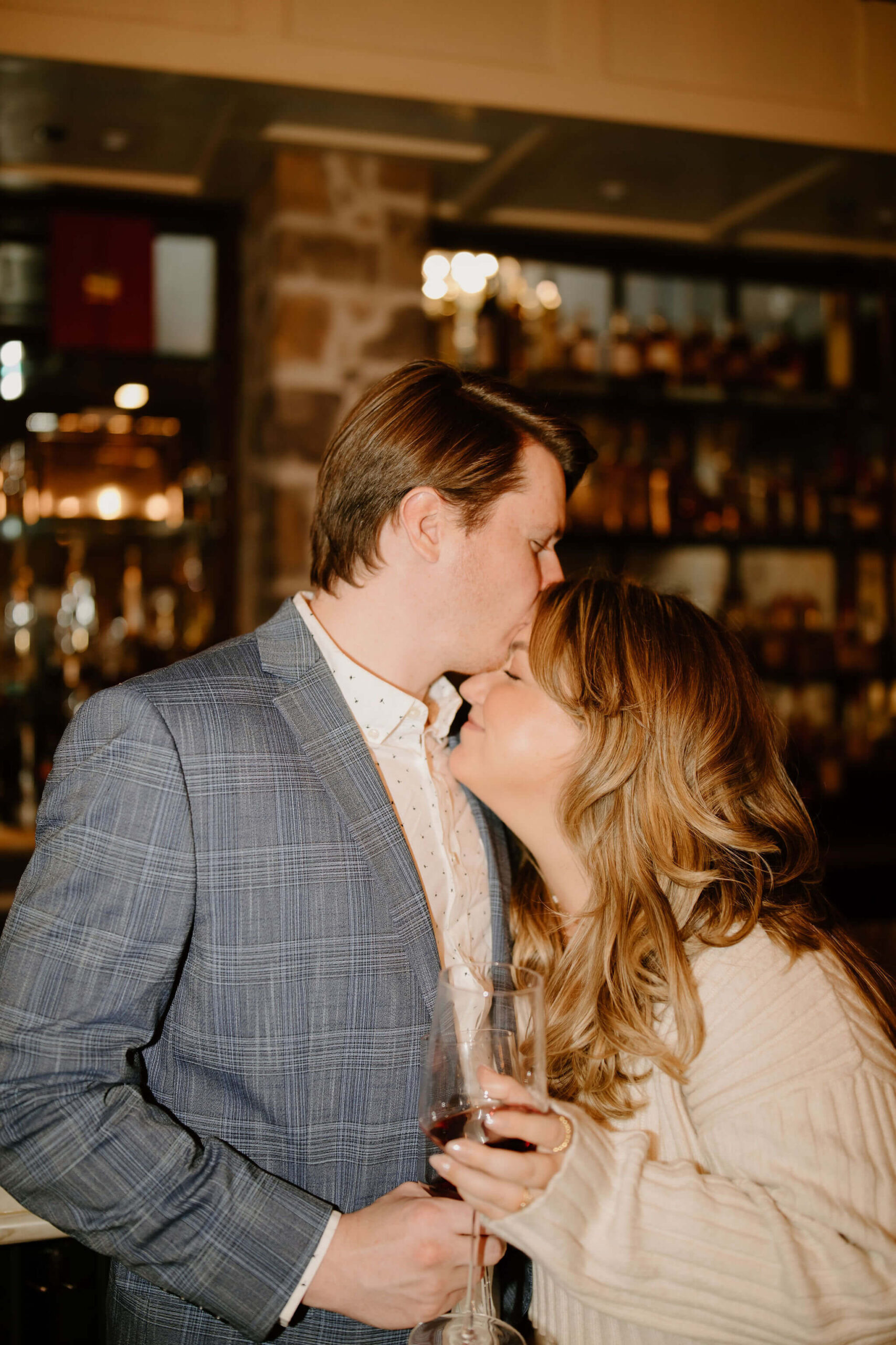 man standing and a woman seated at a bar, both holding wine glasses, while he kisses her on the forehead