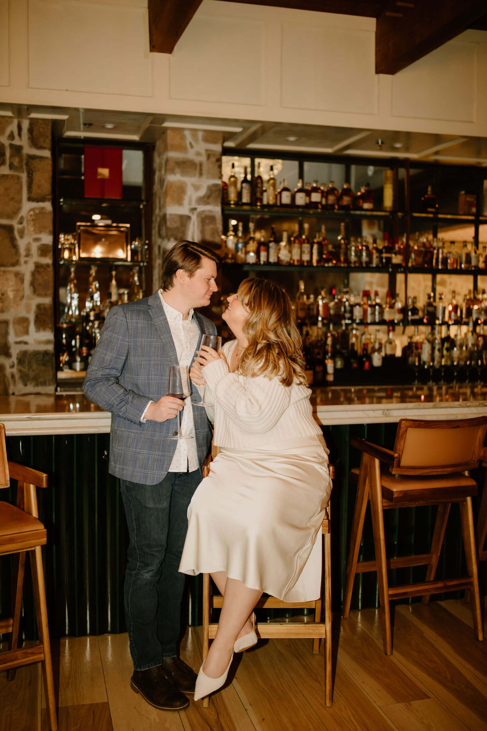 man standing and a woman seated at a bar, both holding wine glasses, leaning into each other and smiling softly at each other