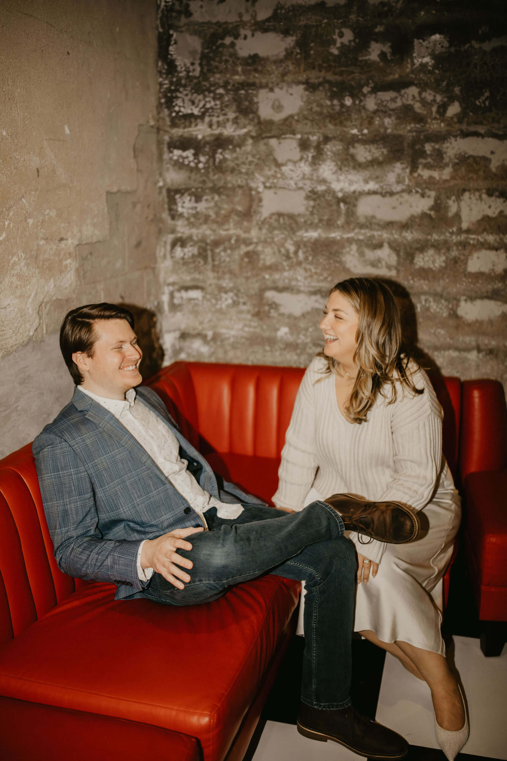 a couple sitting on a red leather couch, leaning toward each other and laughing