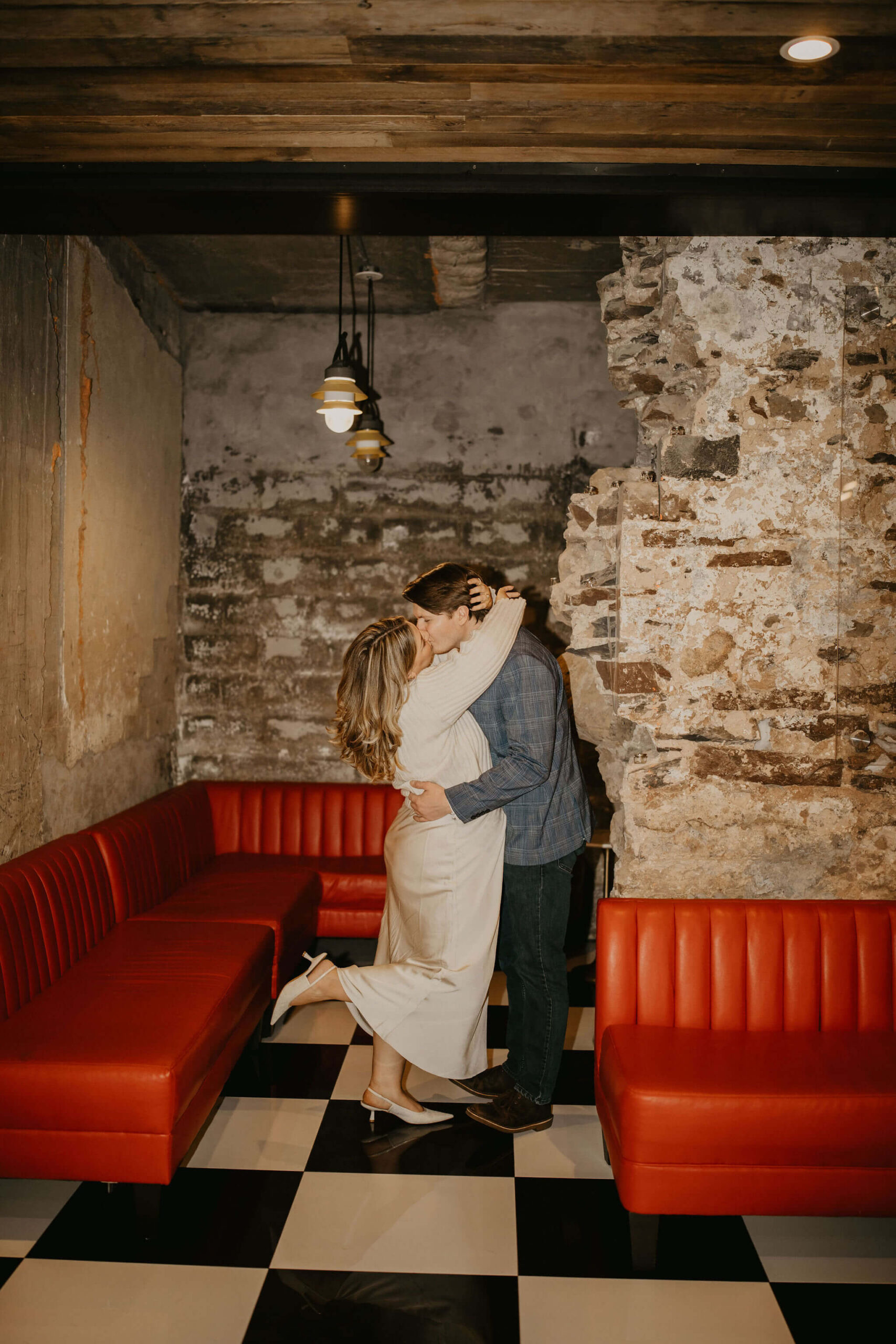 a couple kissing, with her arms around his neck and his arms around her waist. Her one heel is kicked up, and they stand against a stone wall with a red leather couch behind them.