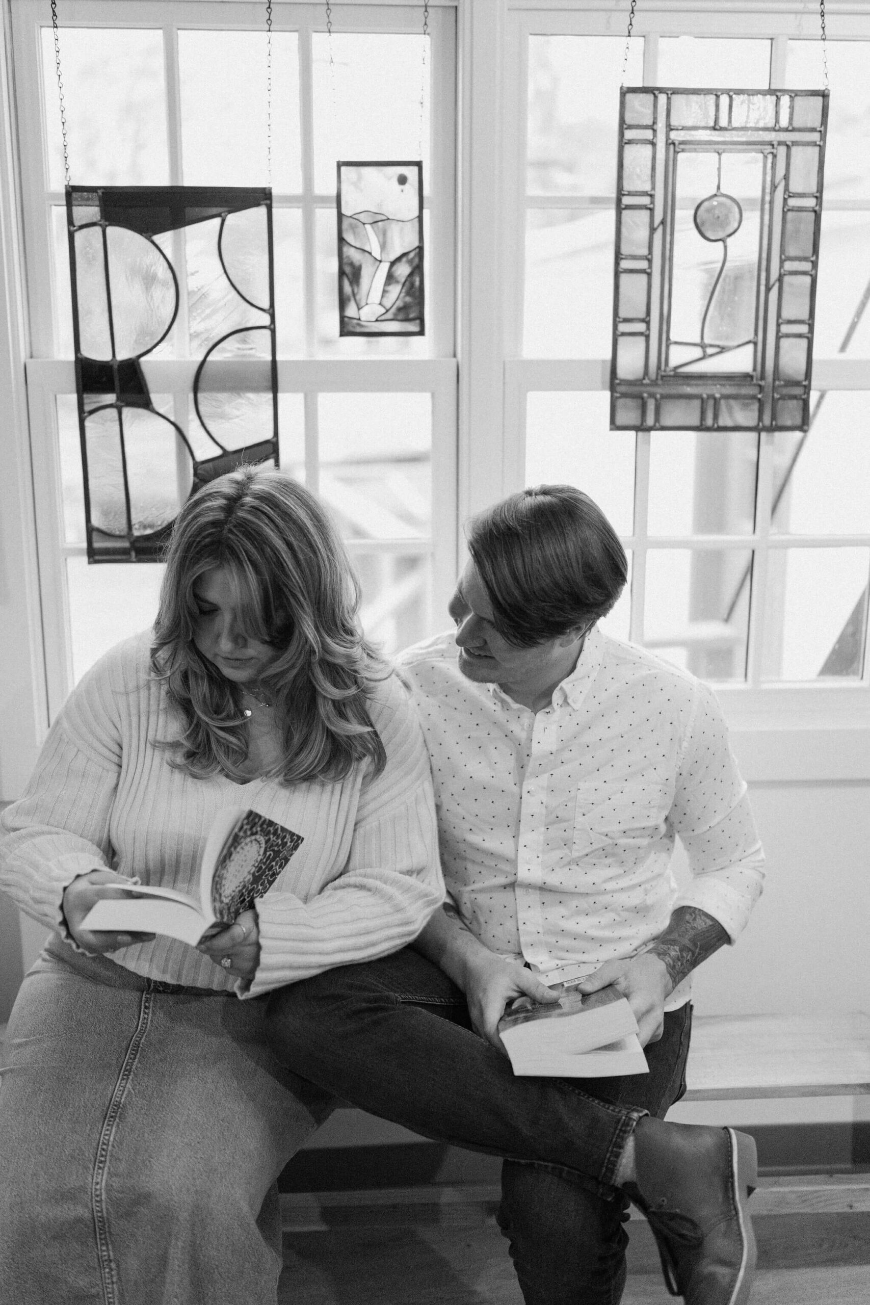 black and white image of a couple sitting on a bench against a window, her reading a book and him reading over her shoulder, while holding his own book