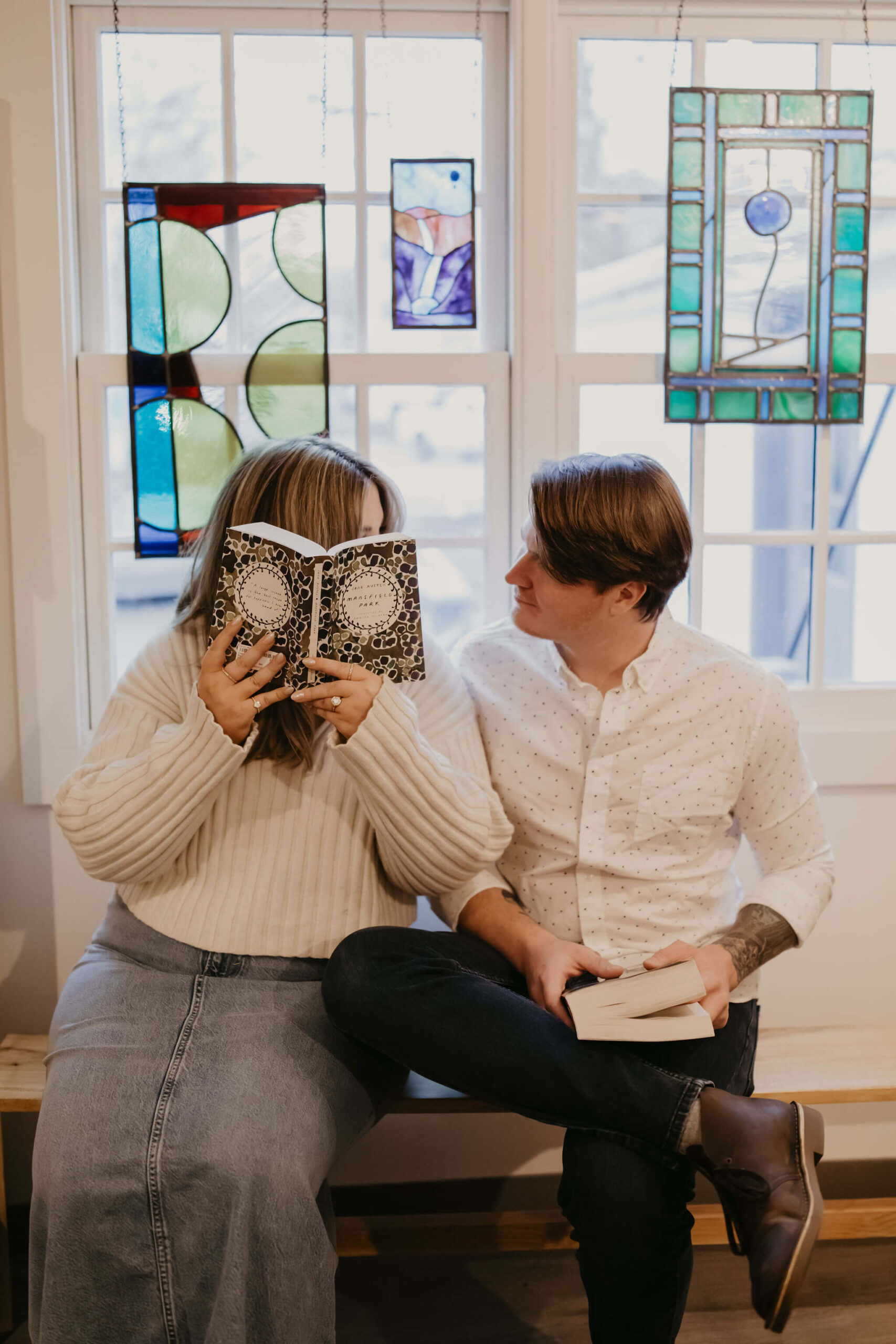 a couple sitting on a bench against a window, her holding a book open covering her face, while he holds a book in his lap looking at her