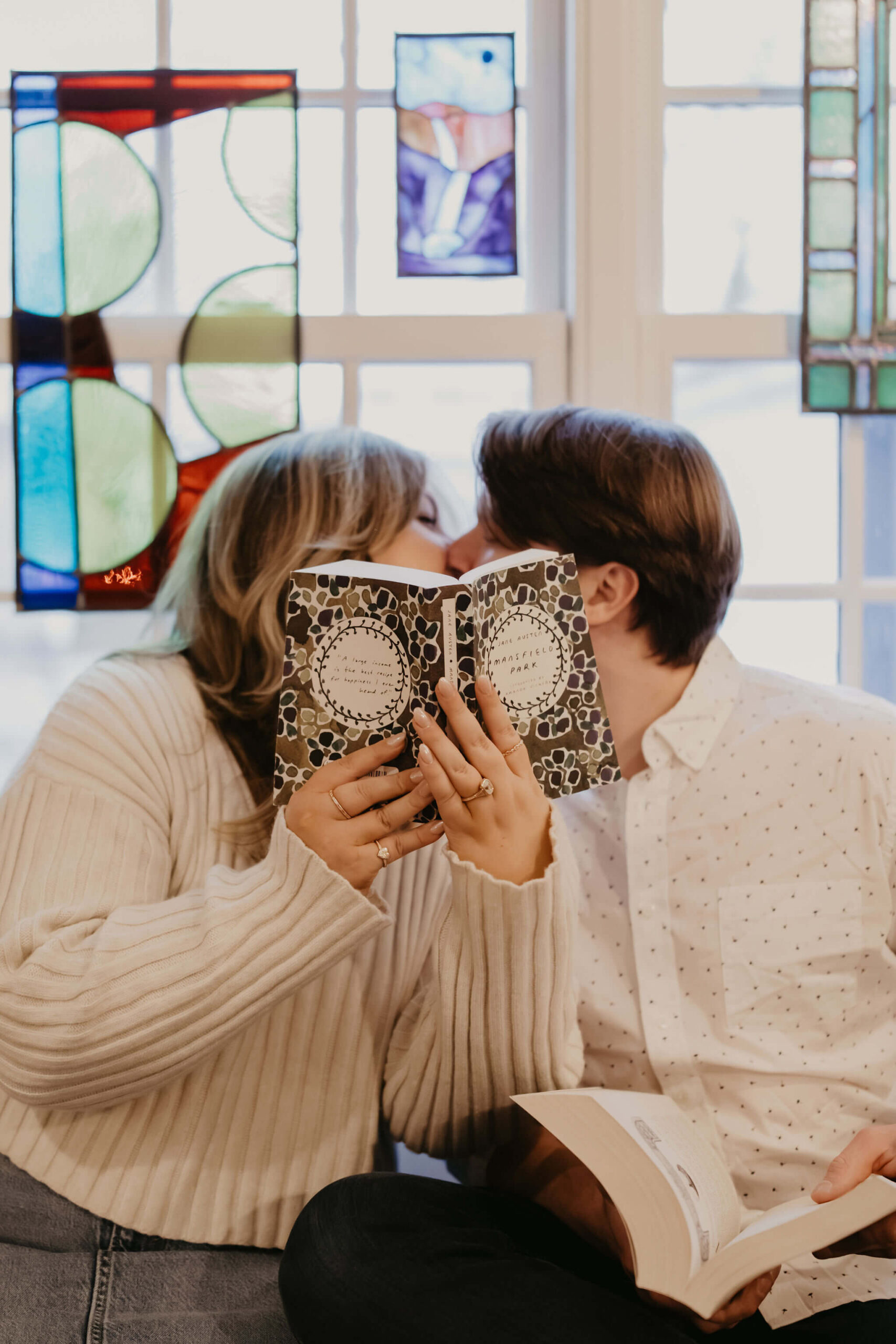 a couple sitting against a stained glass window, kissing behind an opened book