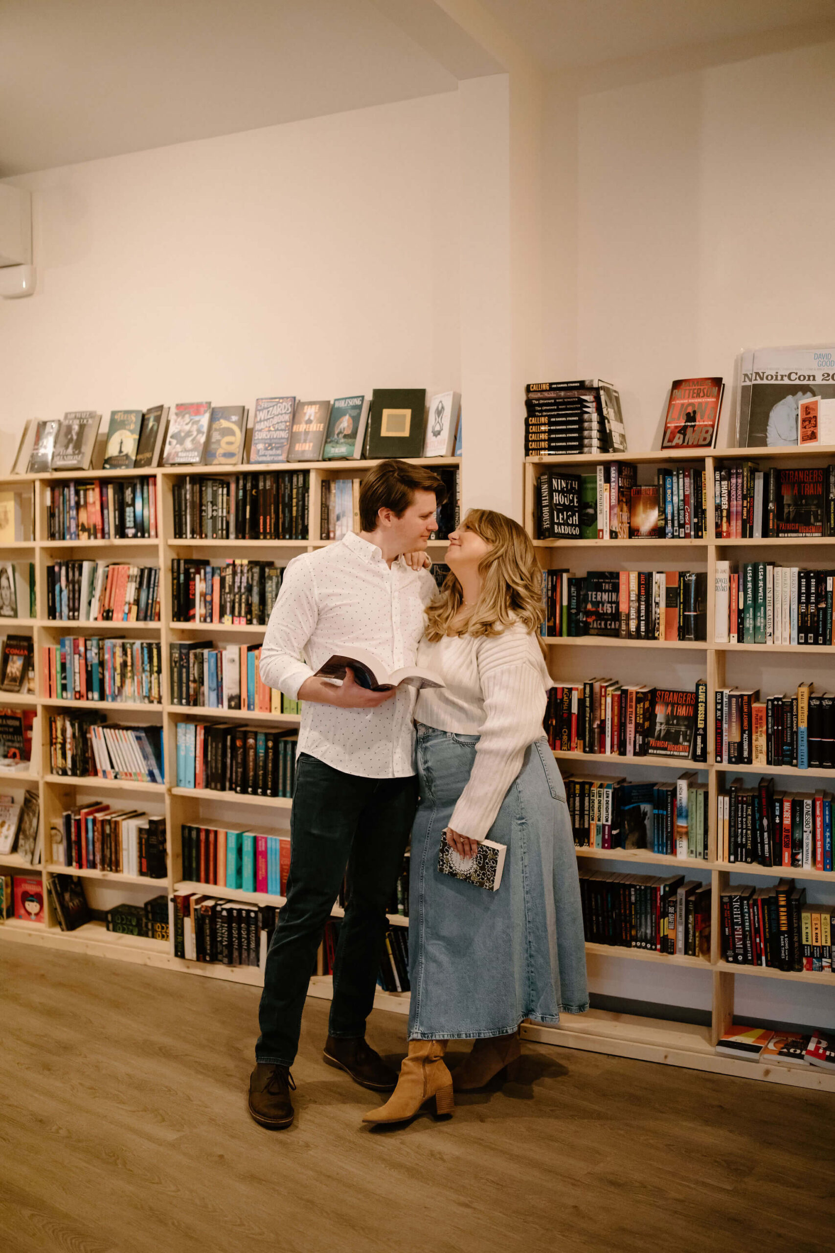 a couple gazing lovingly into each others eyes while standing against a bookshelf, each holding their own books