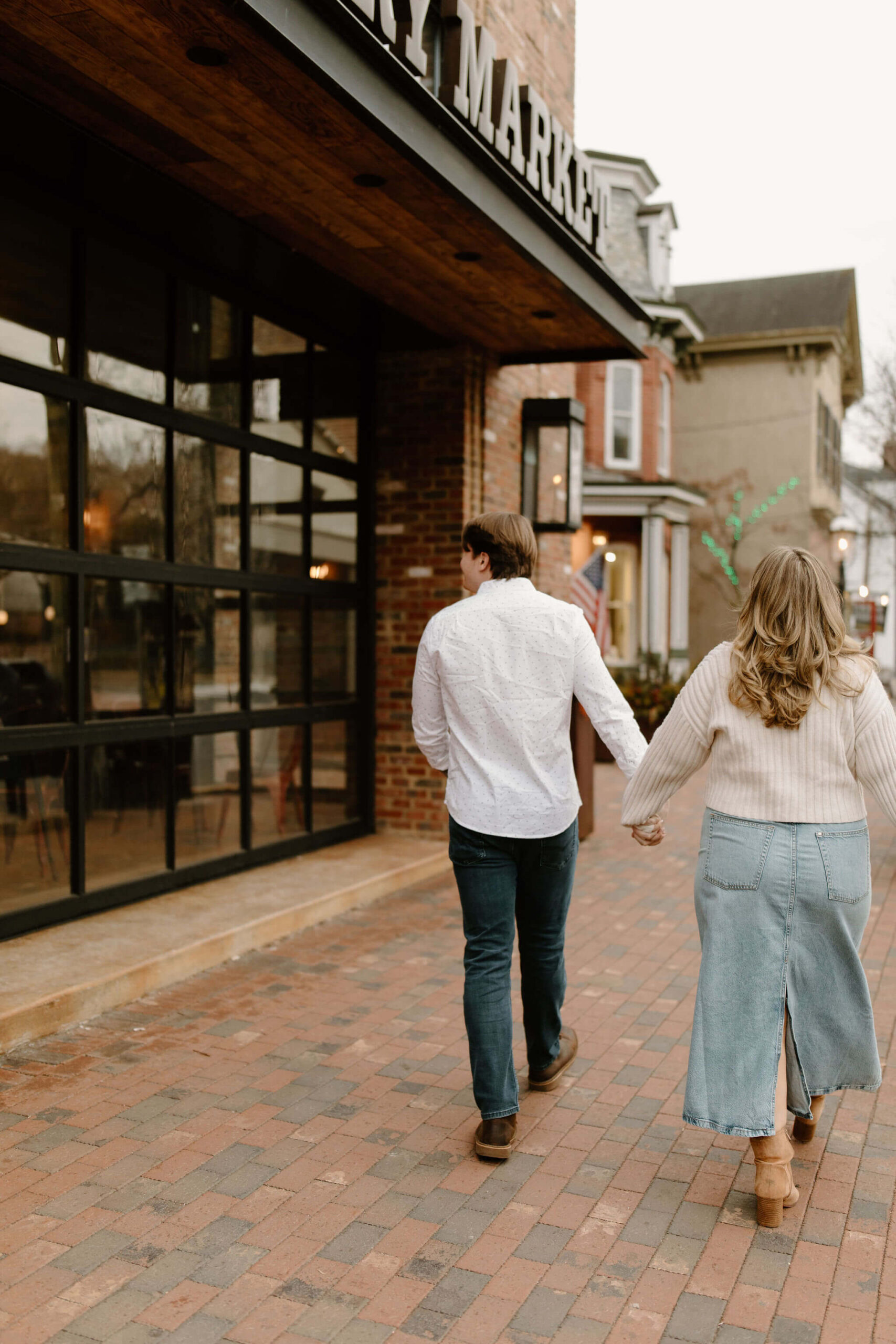 man and a woman holding hands and walking down a brick sidewalk away from the camera