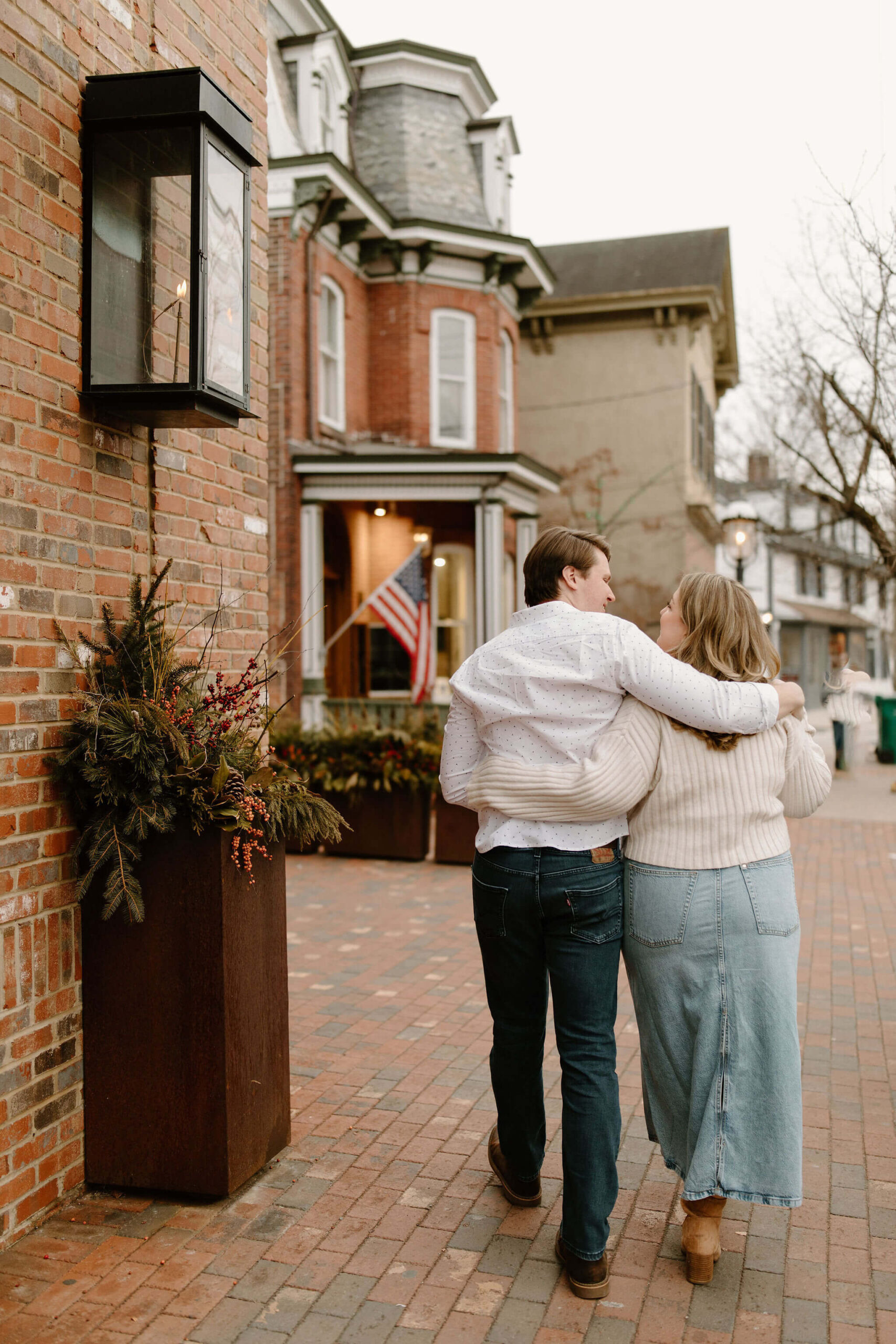 a couple with their arms around each other walking away from the camera down a brick street in a town