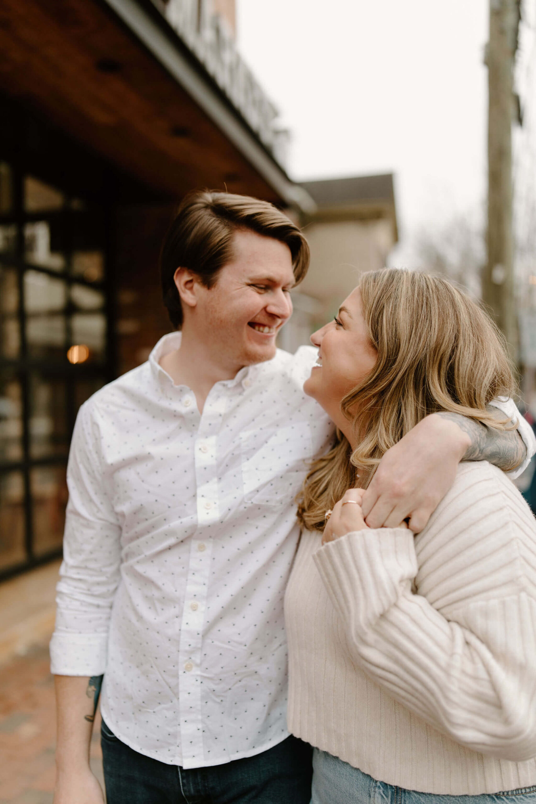 an engaged couple walking toward the camera with his arm around her shoulders and her left hand (with an engagement ring), looking at each other and smiling