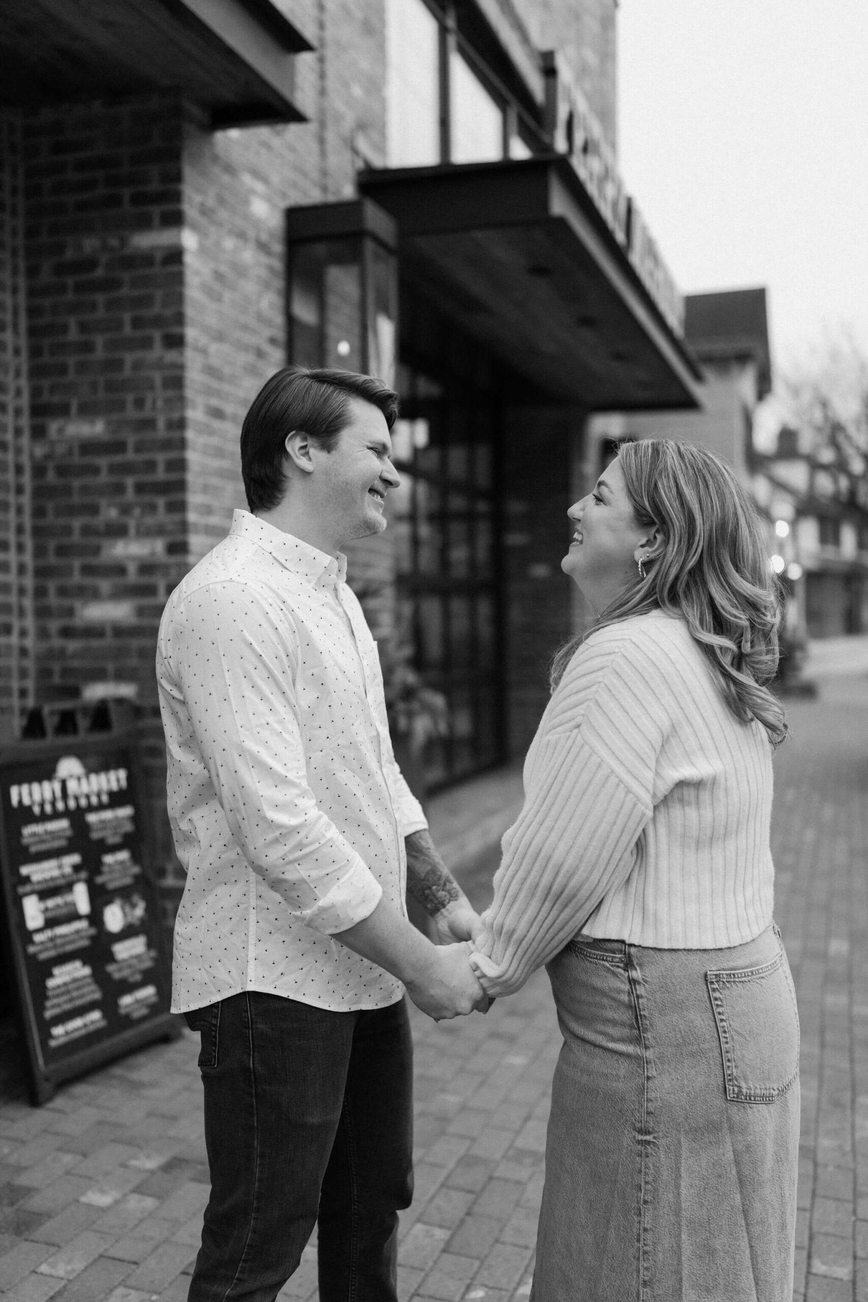 black and white image of a man and a woman holding hands and laughing toward each other outside on a brick sidewalk