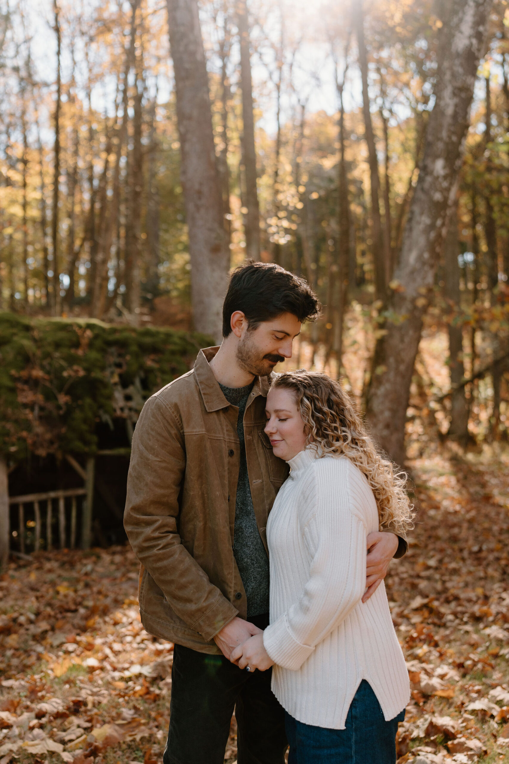 engagement photo of a man and woman holding hands and embracing, leaning into each other with closed eyes in a field surrounded by fall leaves