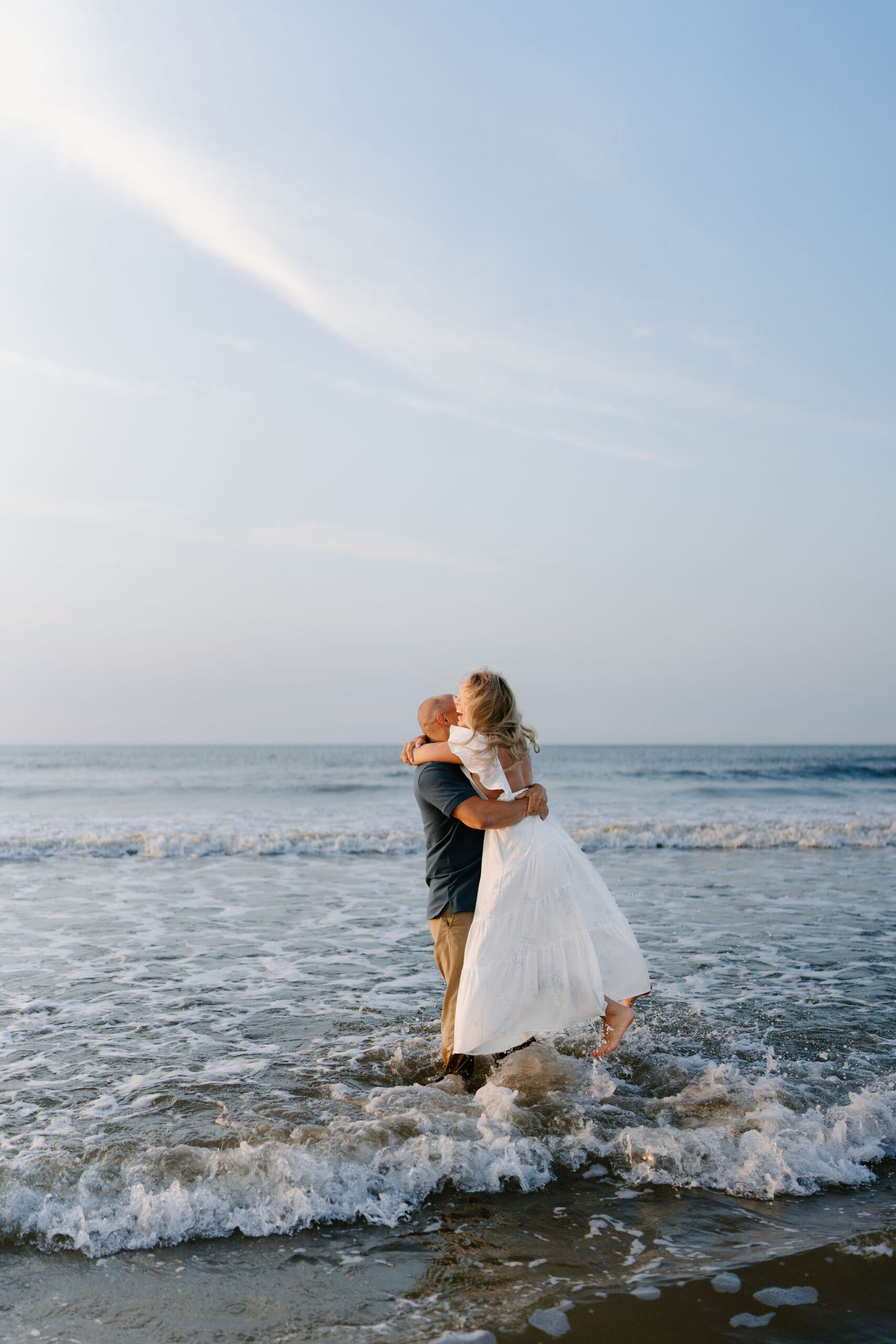 engagement picture of a man (blue shirt, khakis) standing in the ocean while picking his fiance up and spinning her around (wearing a long white dress with an open back), both laughing