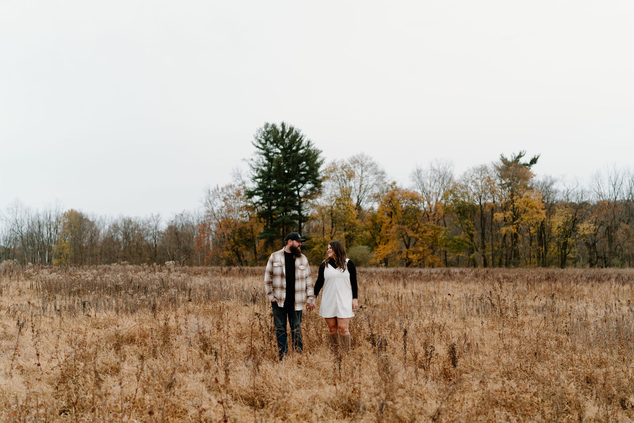 engagement photos of a man with a beard, ball cap, and brown and white flannel, holding hands with his brunette fiance in a white jumper and black turtleneck, smiling at each other