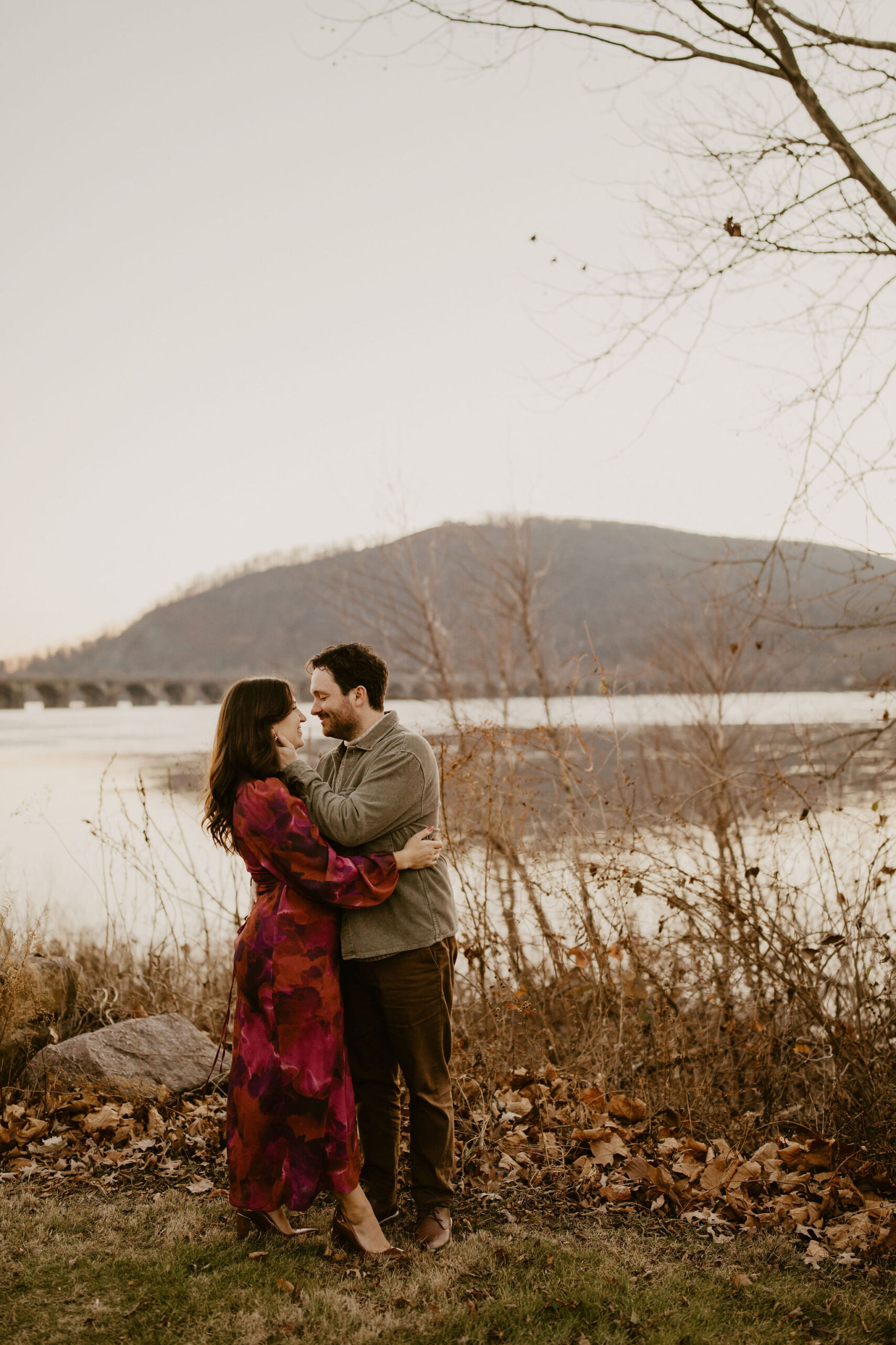 engagement photos of a man holding his fiance's face (wearing a red nad purple patterned dress) and smiling just before kissing her, while standing in front of a river in the winter