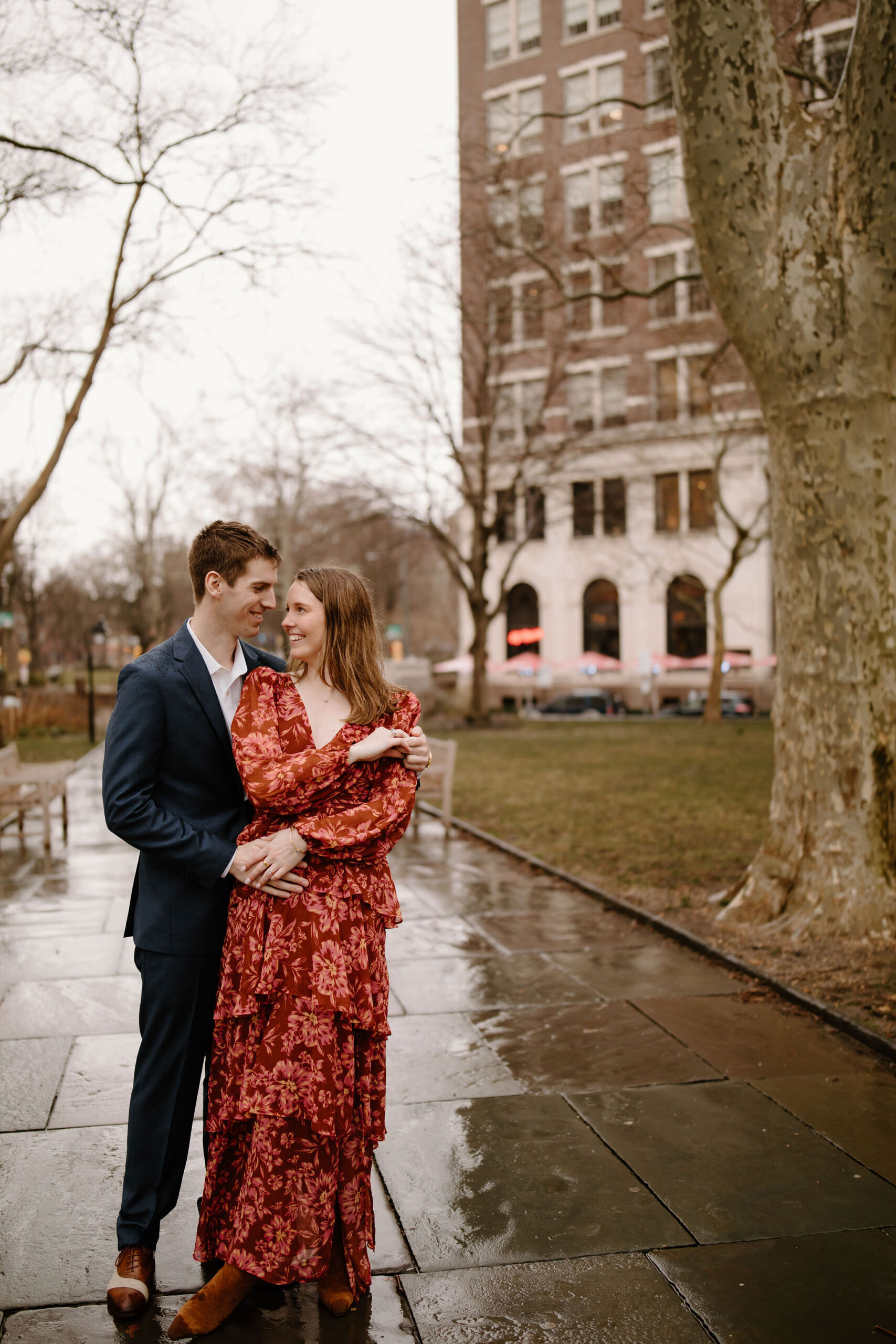 engagement photos of a man in a navy blue suit and brown leather dress shoes, with his arms around his fiance in a red tiered flowy dress, smiling at each other in the city