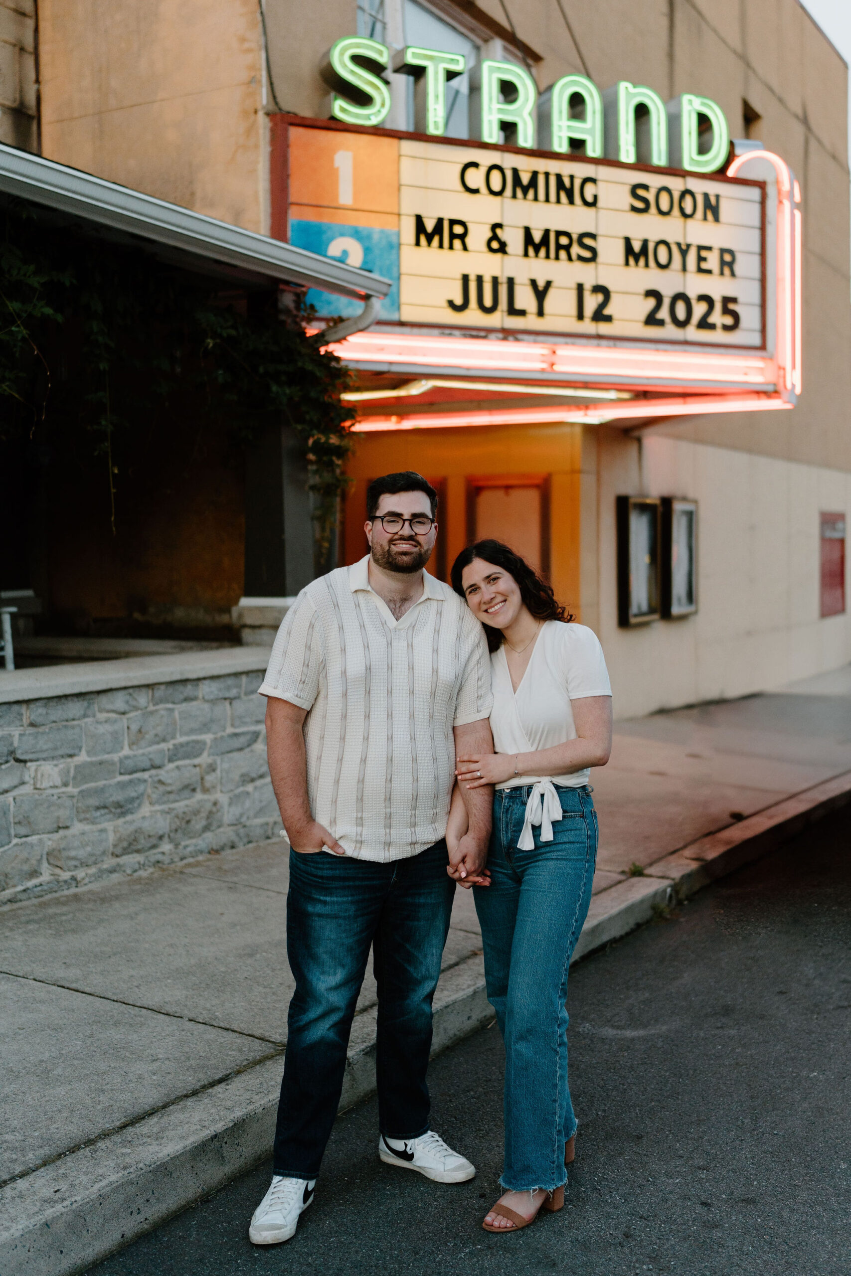 engagement photos of a couple (both  brunette, wearing a white shirt and jeans) smiling at the camera while standing in front of a custom movie theater marquis that reads "coming soon, Mr. & Mrs. Moyer, July 12, 2025"