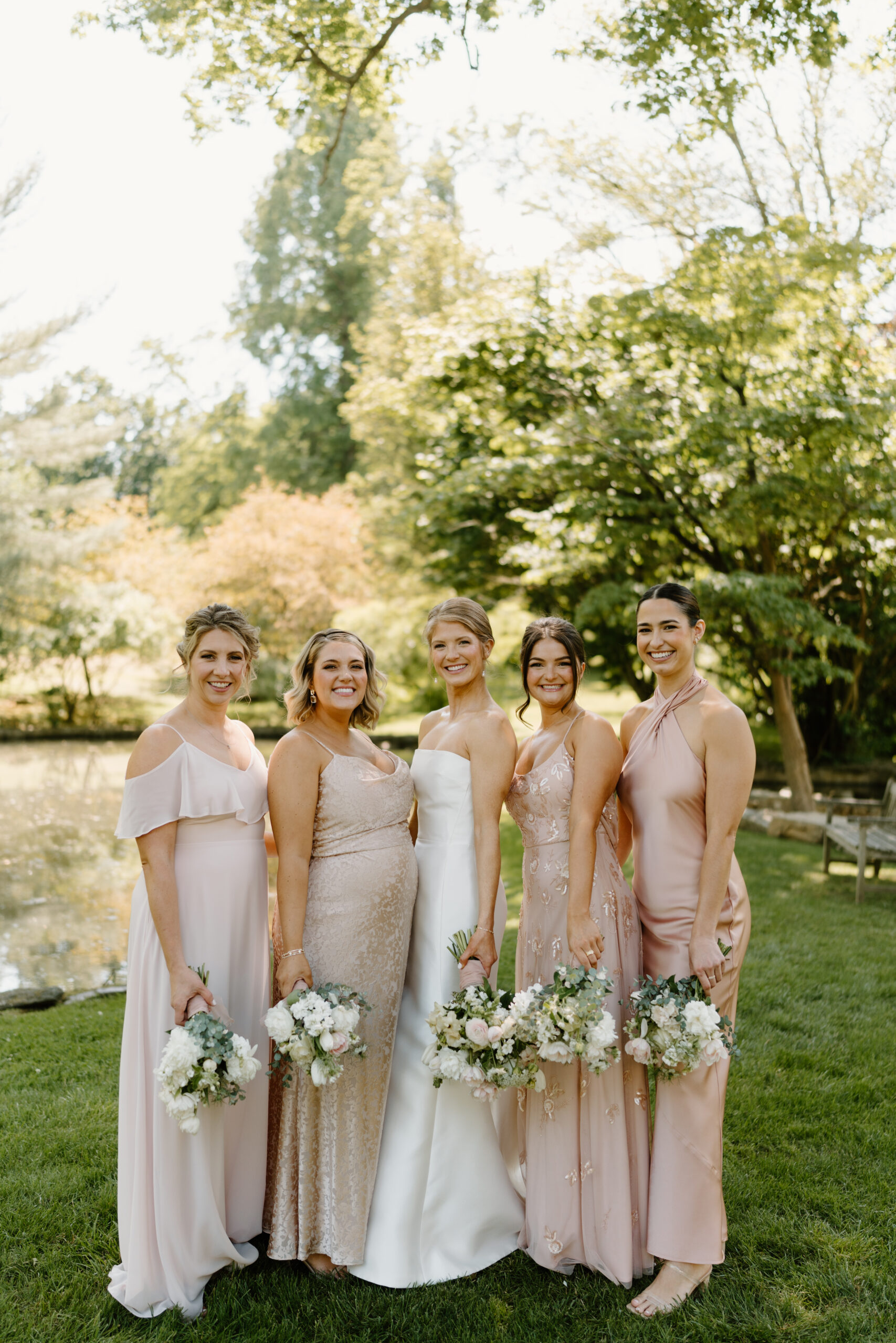 a bride (white dress) and four bridesmaids (light pink dresses) holding flowers at their sides and smiling at the camera