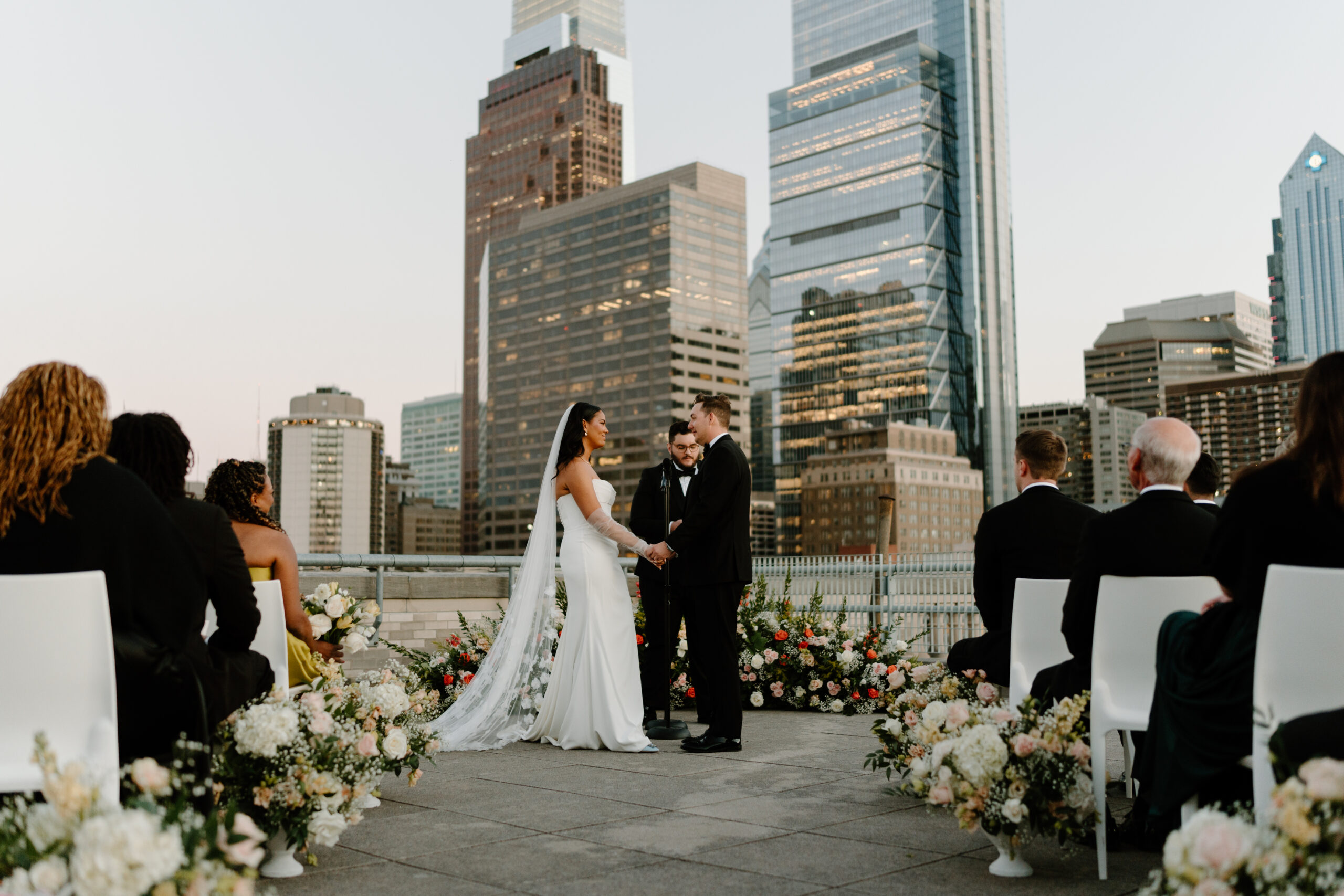 bride and groom holding hands and smiling softly at each other during their rooftop wedding ceremony, with guests sitting in white chairs watching, with the philadelphia skyling behind them