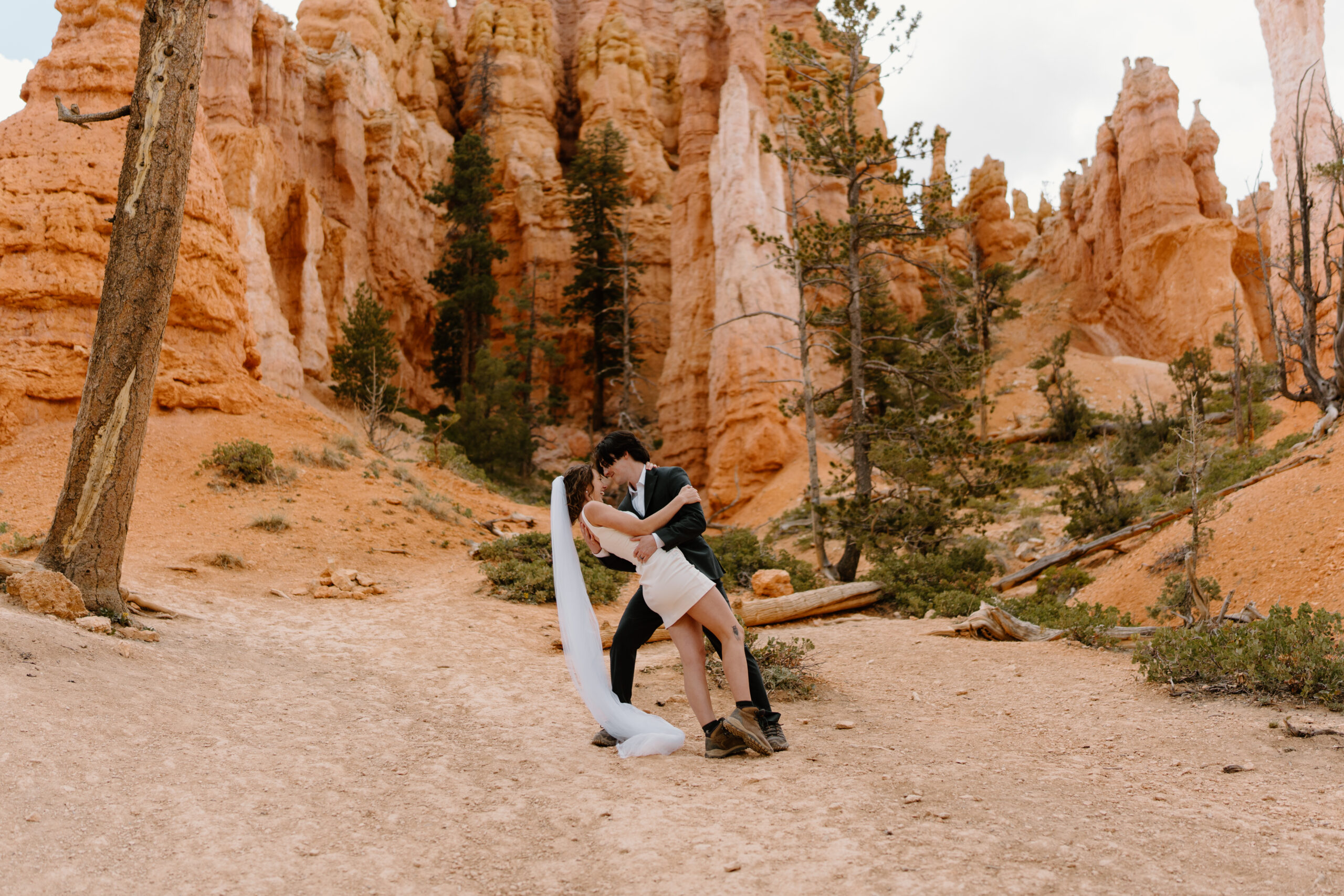 groom (charcoal gray suit, hiking boots) dipping bride (short white dress, long veil, hiking boots) during their elopement at Bryce Canyon National Park while on a hike 