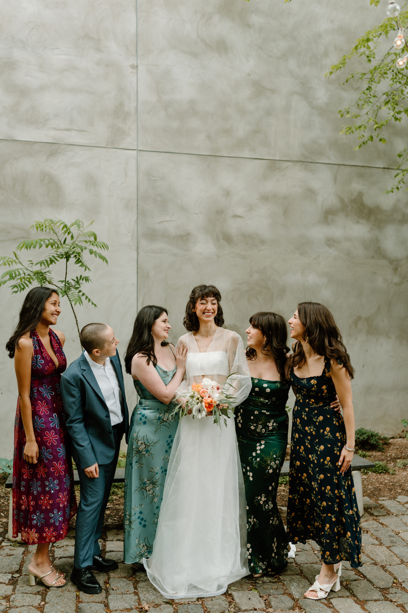 a bride (wearing white) and five friends (four in colorful floral dresses, one in a blue suit) smiling at each other