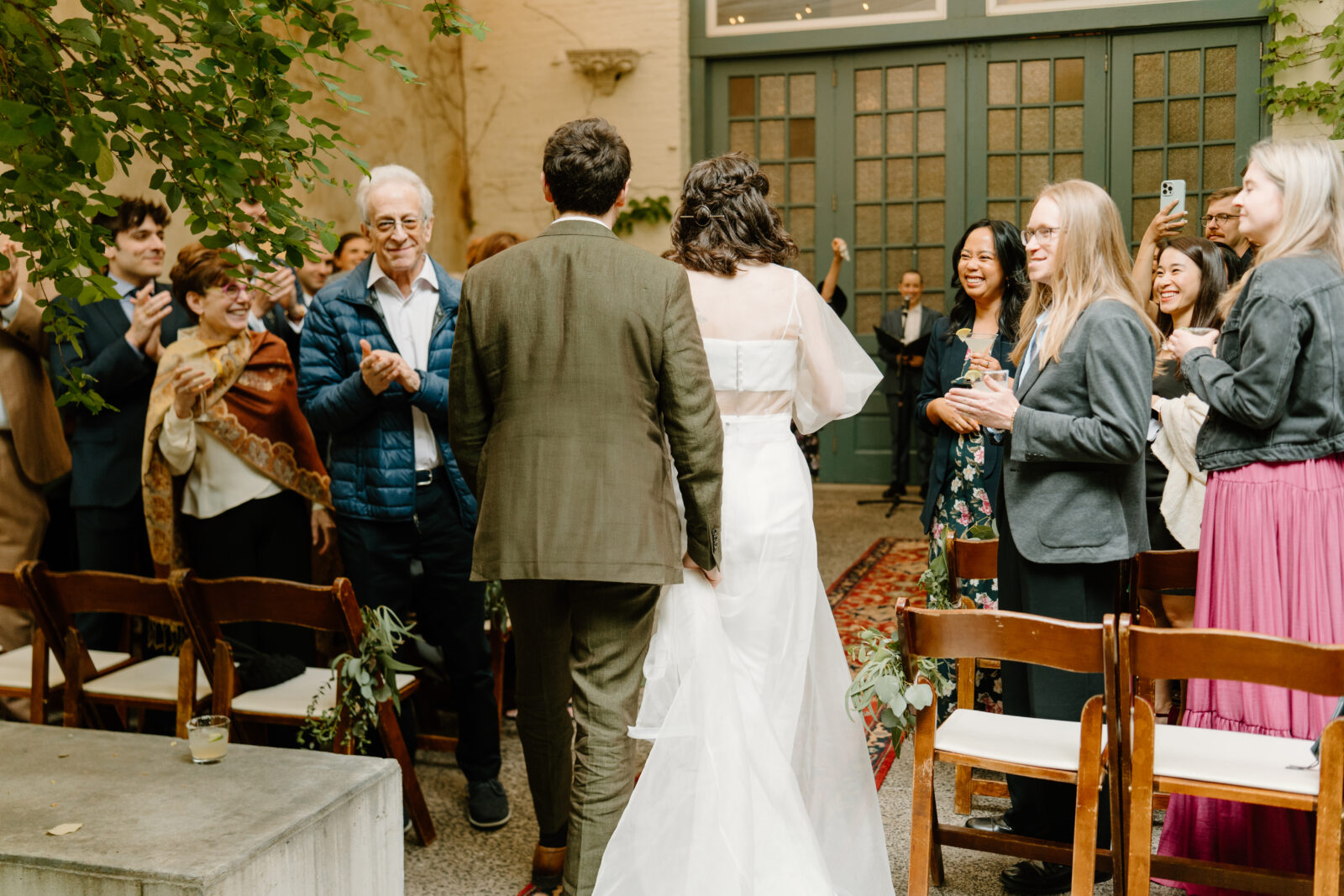 bride (cropped white top and skirt) and groom (green suit) walking down the aisle to enter their ceremony together