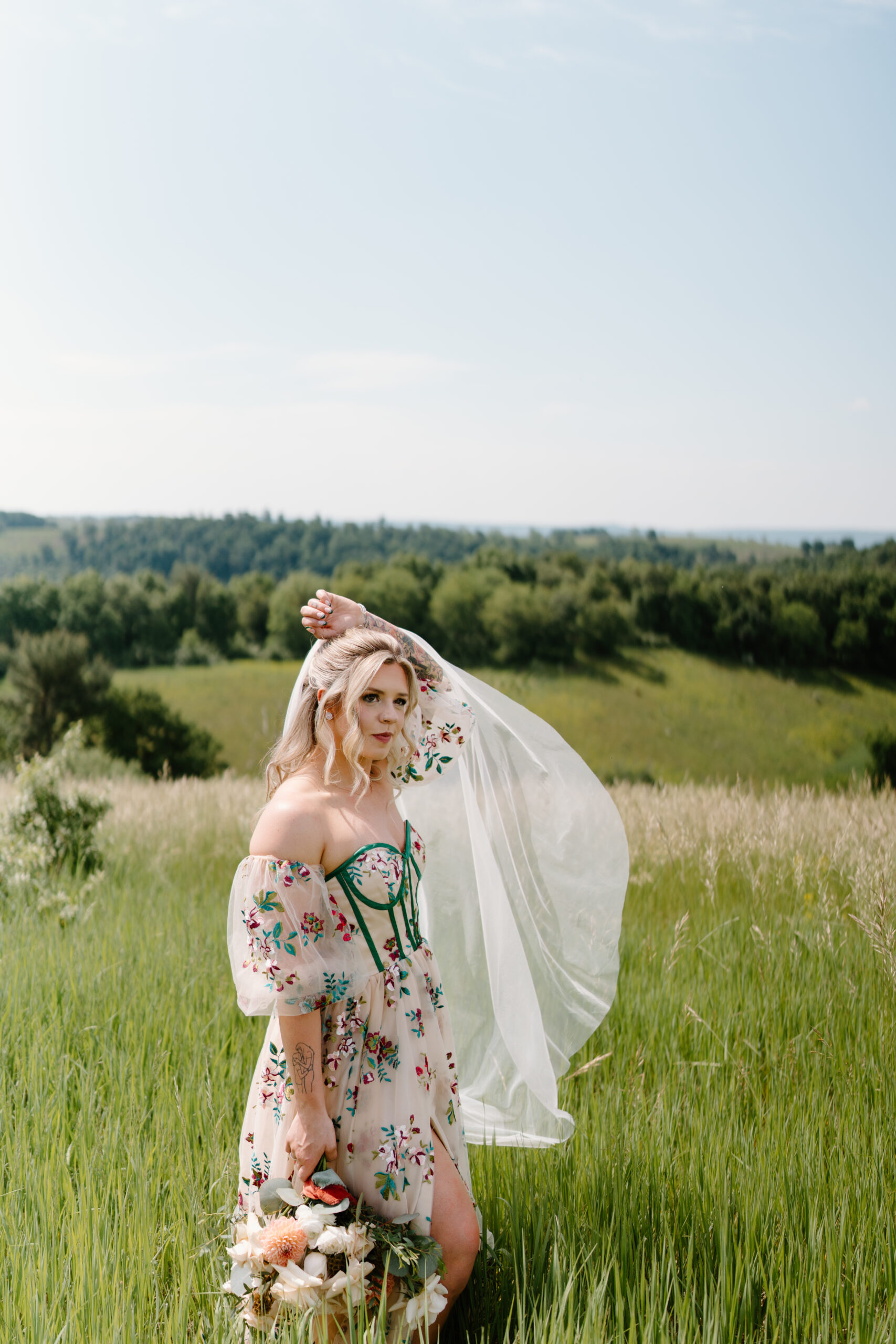 bride with blonde hair tossing her veil in the breeze and holding her tattooed left arm over her head, smiling softly off camera and holding her bouquet in her right hand at her side. her wedding dress is pale pink with colorful embroidered flowers and green corset boning.