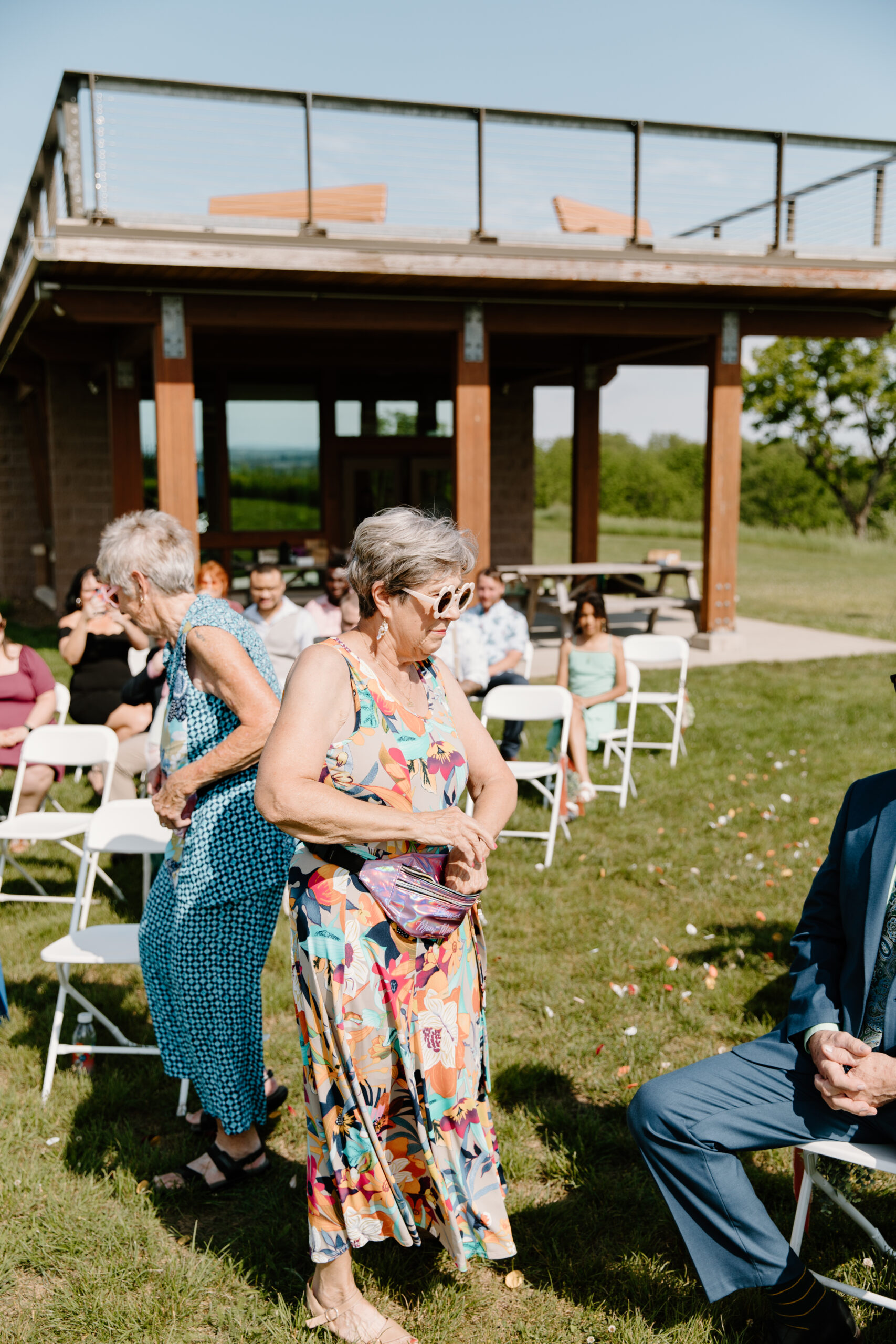 two grandmothers in colorful dresses, serving as the flower girls for a wedding ceremony, wearing sunglasses and throwing flower petals out of the fanny packs they wear