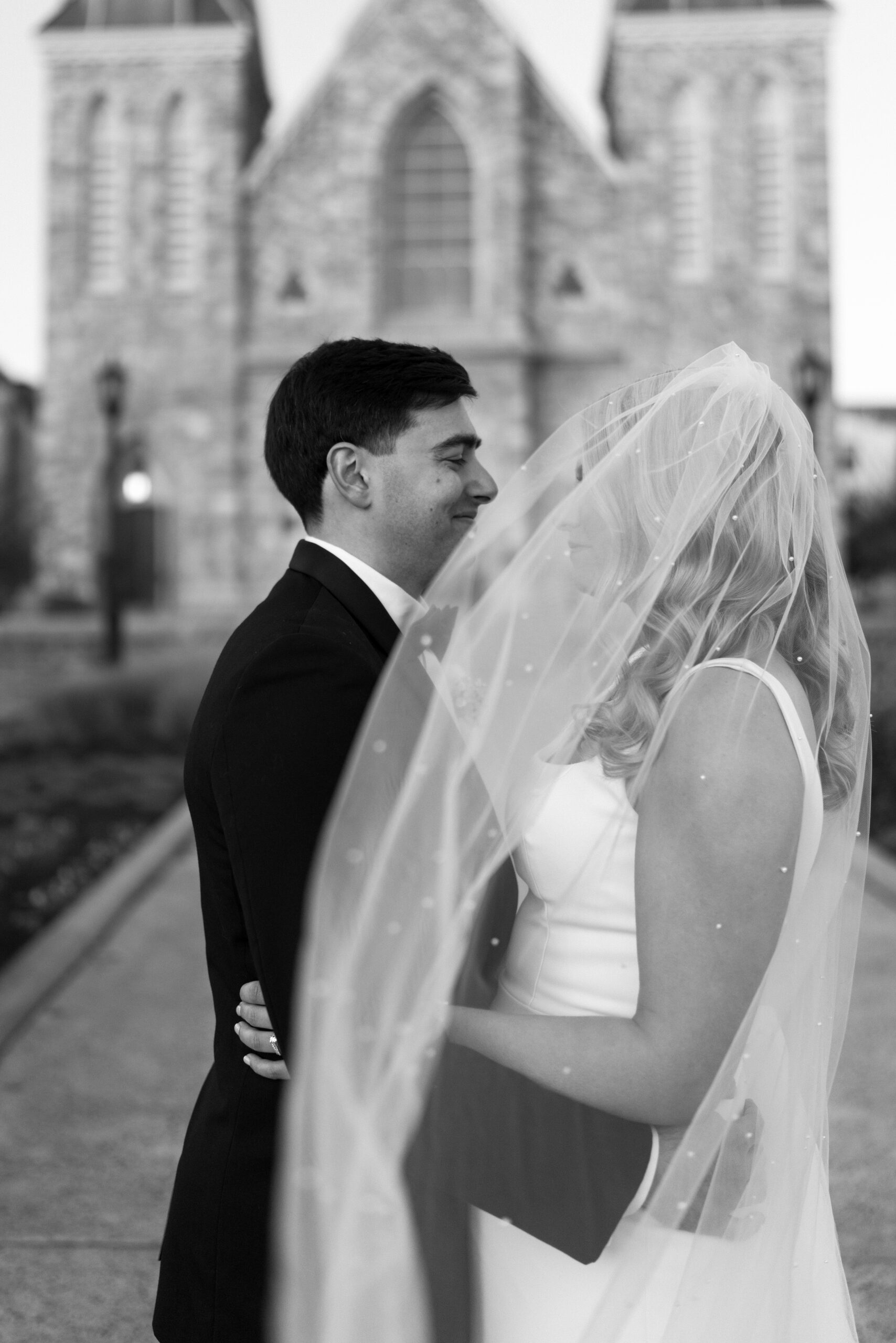 black and white image of groom (black suit) and bride (blonde hair, white dress) embracing and smiling at each other while her veil blows in front of them