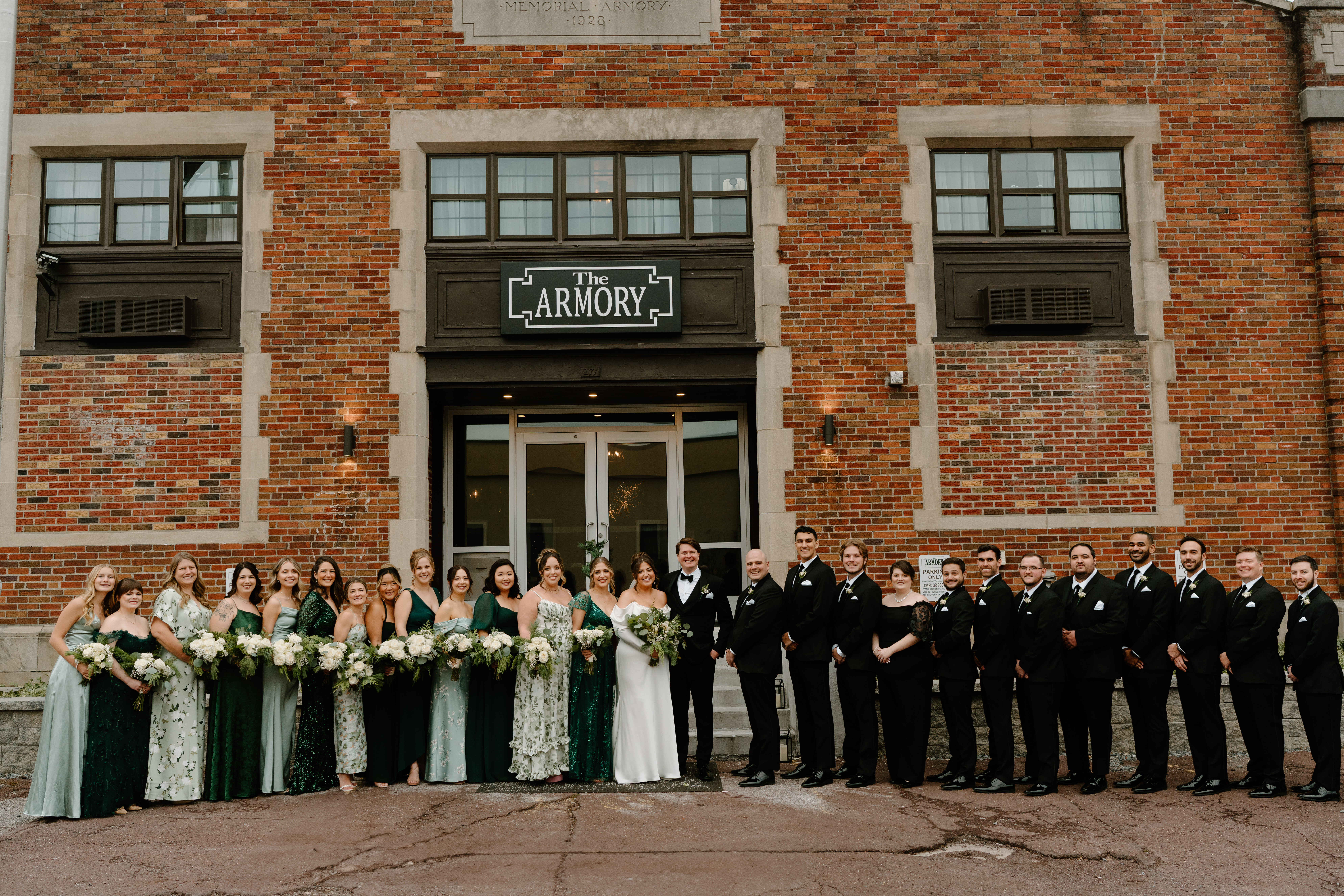 a large wedding party portrait of mixed genders, the bride's side wearing green patterned dresses and the groom's side wearing all black dresses and suits. All are smiling at the camera in front of a brick building.