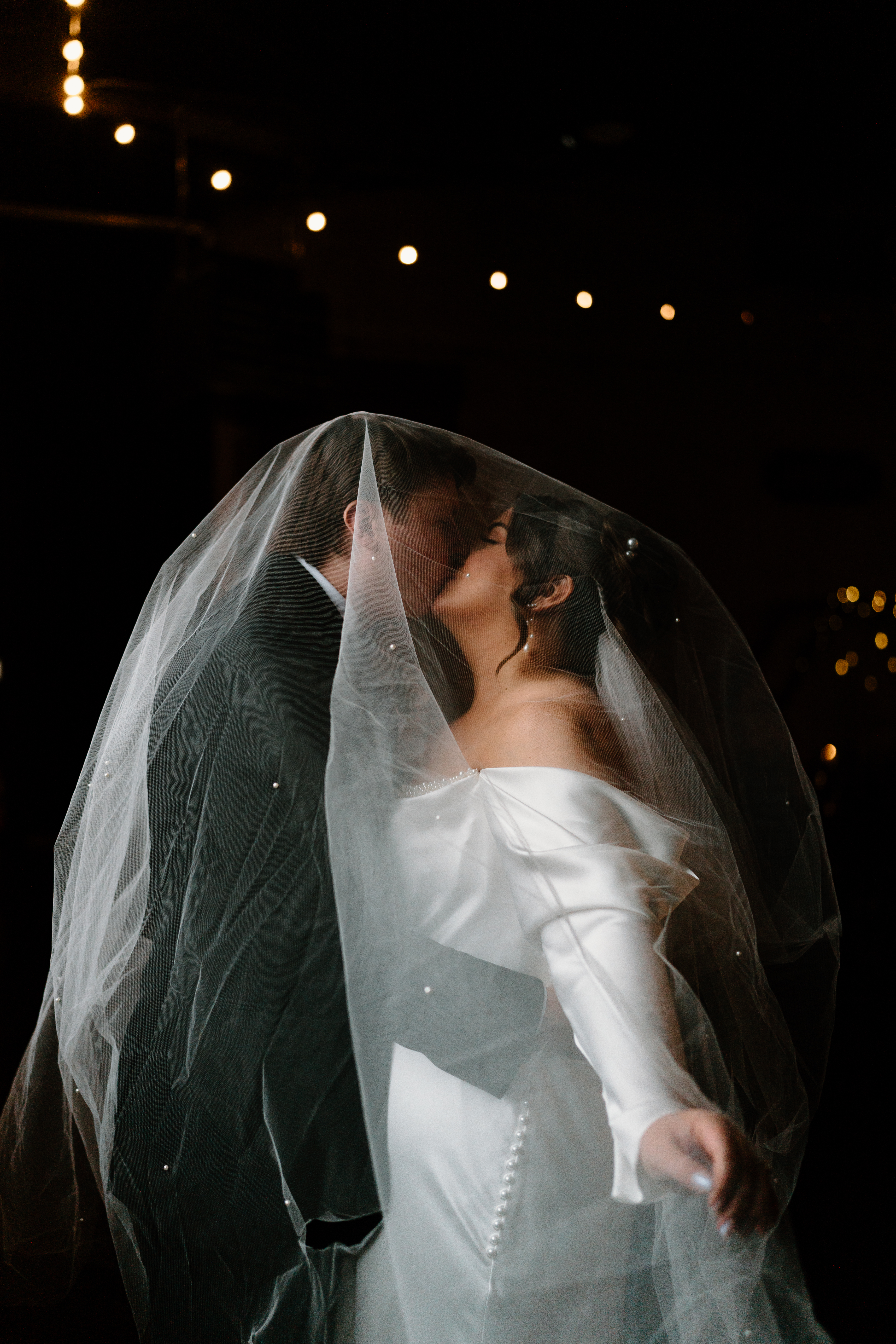 bride (brown hair, long sleeved white dress) and groom (light brown hair, black suit) kissing underneath a wedding veil with inlayed pearls