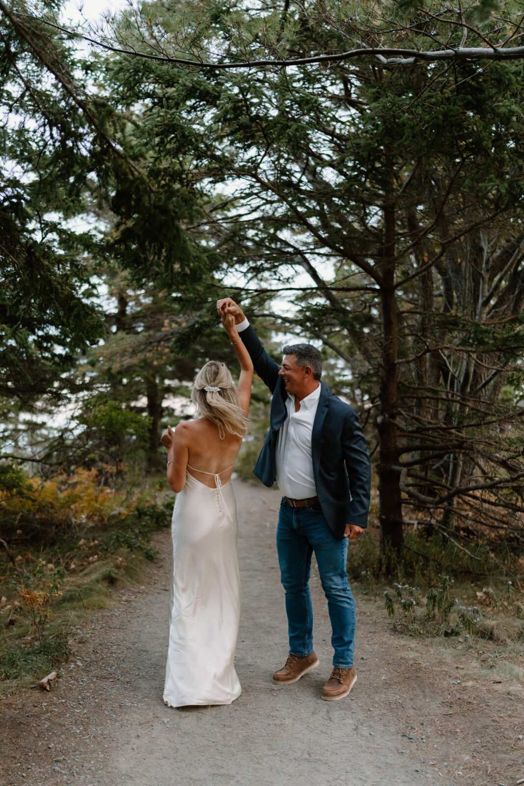 a man (jeans, white shirt, navy blue blazer) twirling his wife (blonde hair, white backless sleek wedding dress) and smiling, on a dirt trail surrounded by evergreen trees