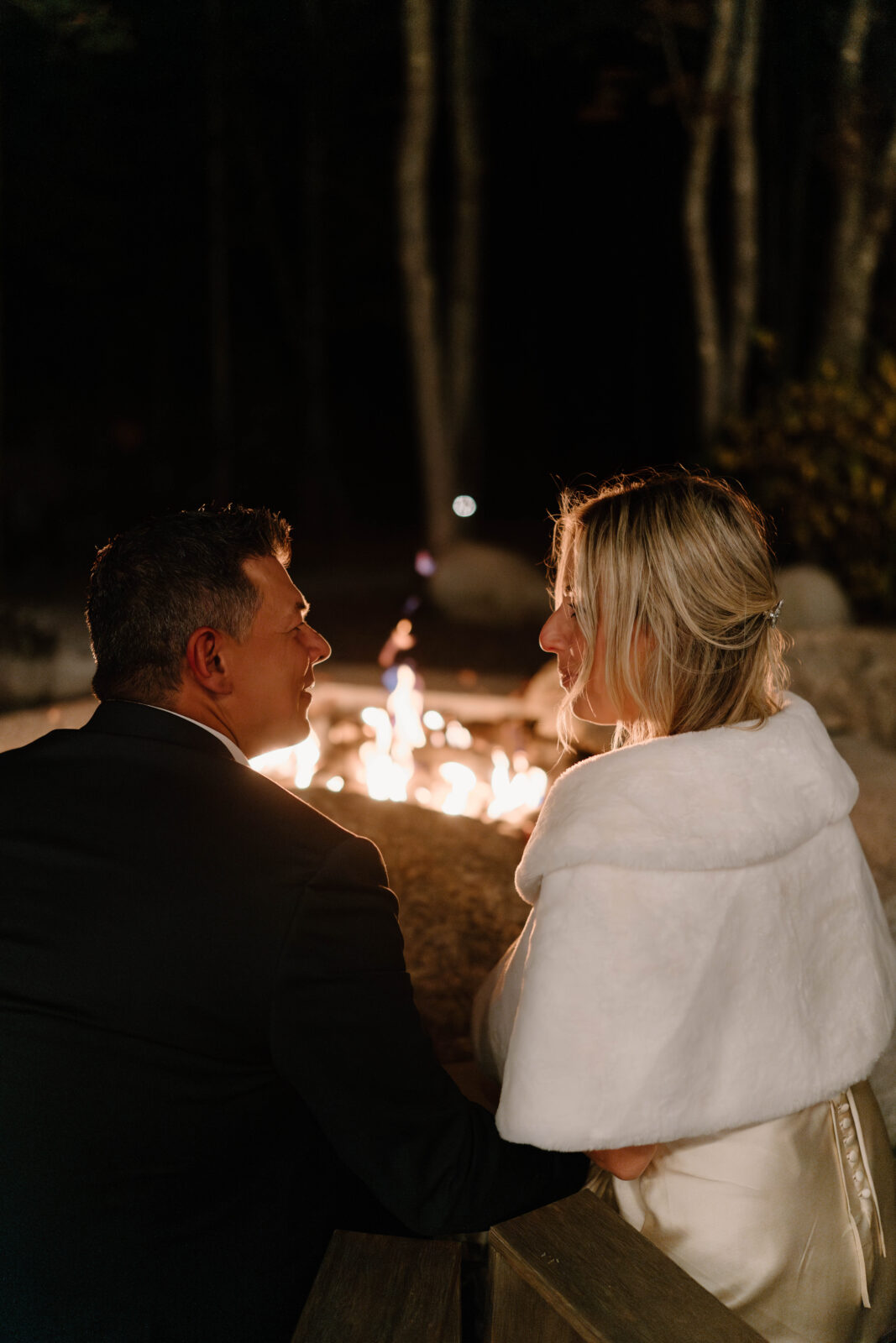 close up image of a bride (blonde, in a white dress and a furry white shaw) and groom (salt and pepper hair, dark blue suit jacket) smiling at each other while sitting in front of a bonfire