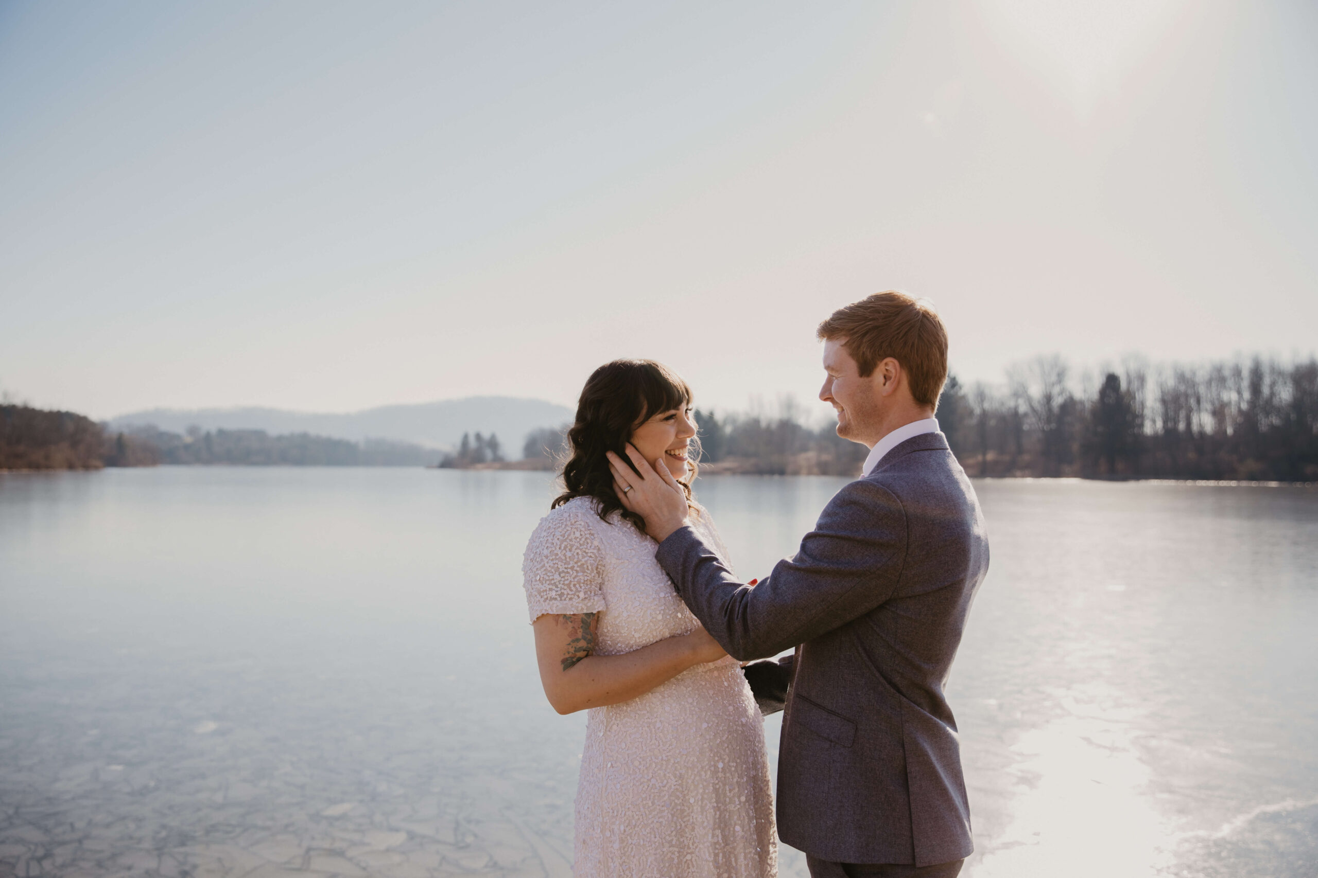 bride (brown hair with bangs and a sequined white dress) smiling at her groom (gray suit, red hair) who is holding her face with his left hand, about to kiss her. They are standing in front of a frozen lake in bright sun, with a mountain in the distance